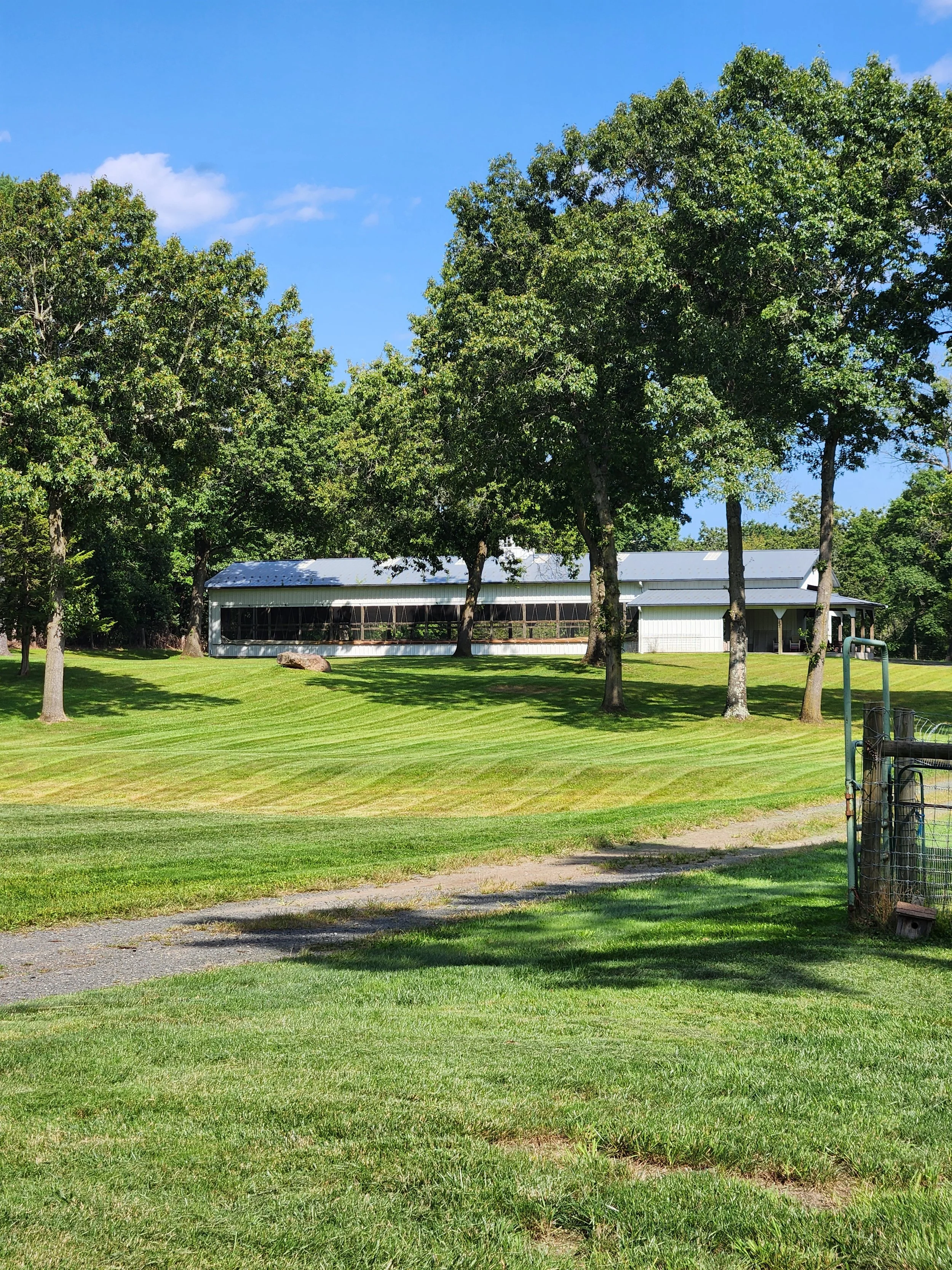 A large, light-colored building with a metal roof, situated in a lush, green landscape with several tall trees, grass with mowing patterns, and a blue sky with some clouds.