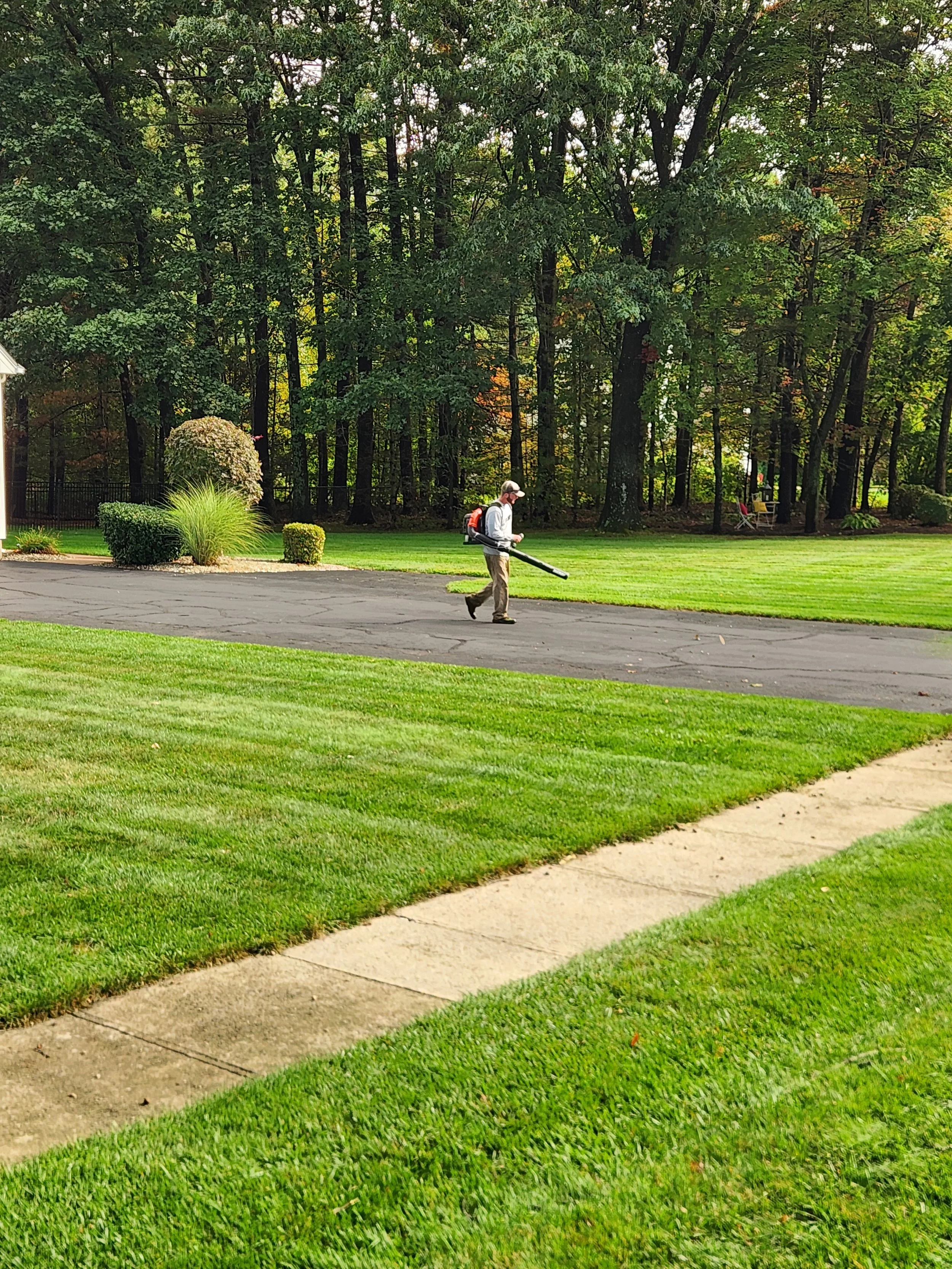 A person wearing a hat, gray long-sleeve shirt, khaki pants, and black shoes is walking on a driveway, holding a leaf blower. The scene is outdoors with green grass, trees, and shrubs in the background.