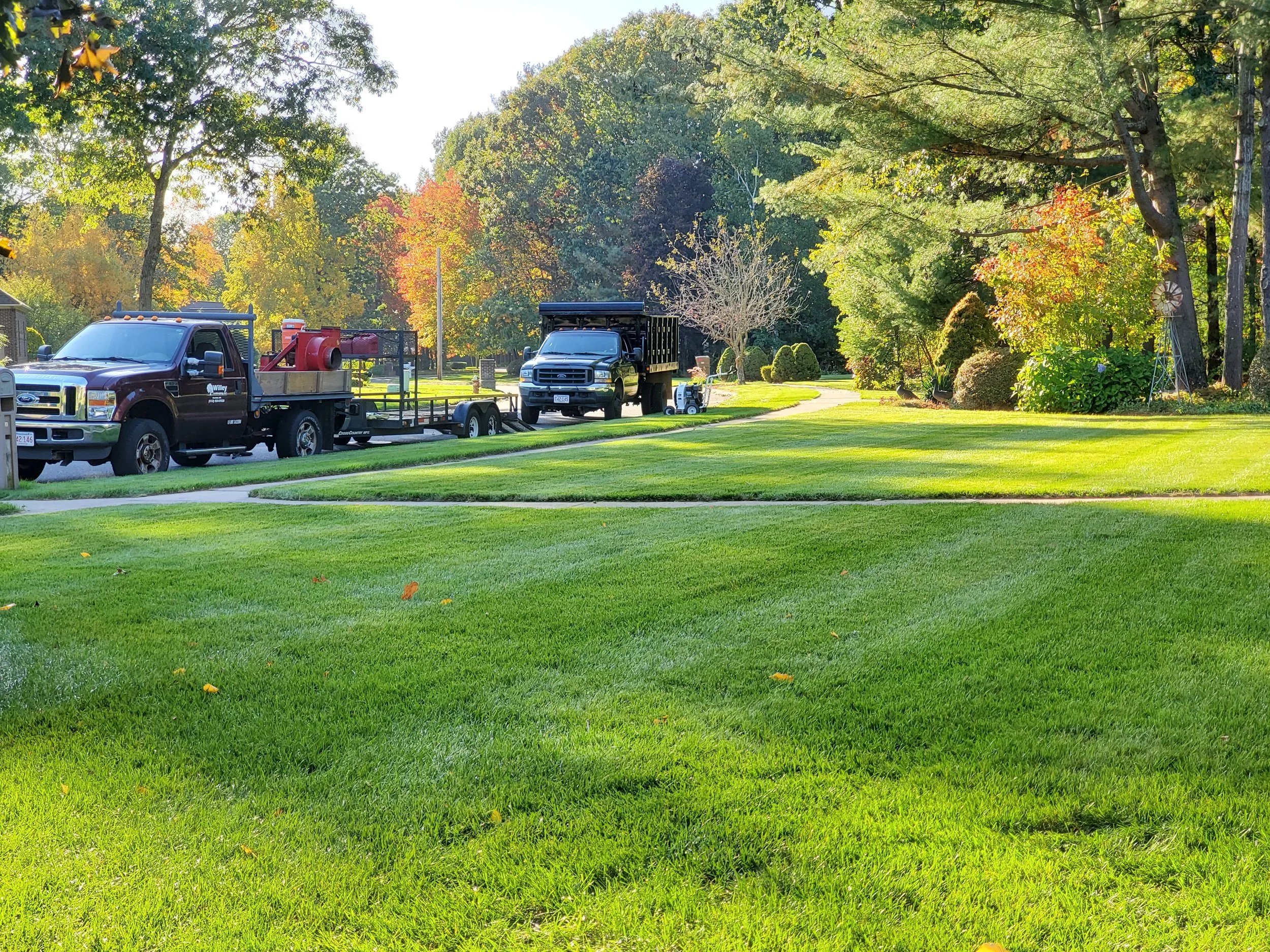 Green lawn with two trucks parked on the street, houses, and trees with fall foliage in the background.