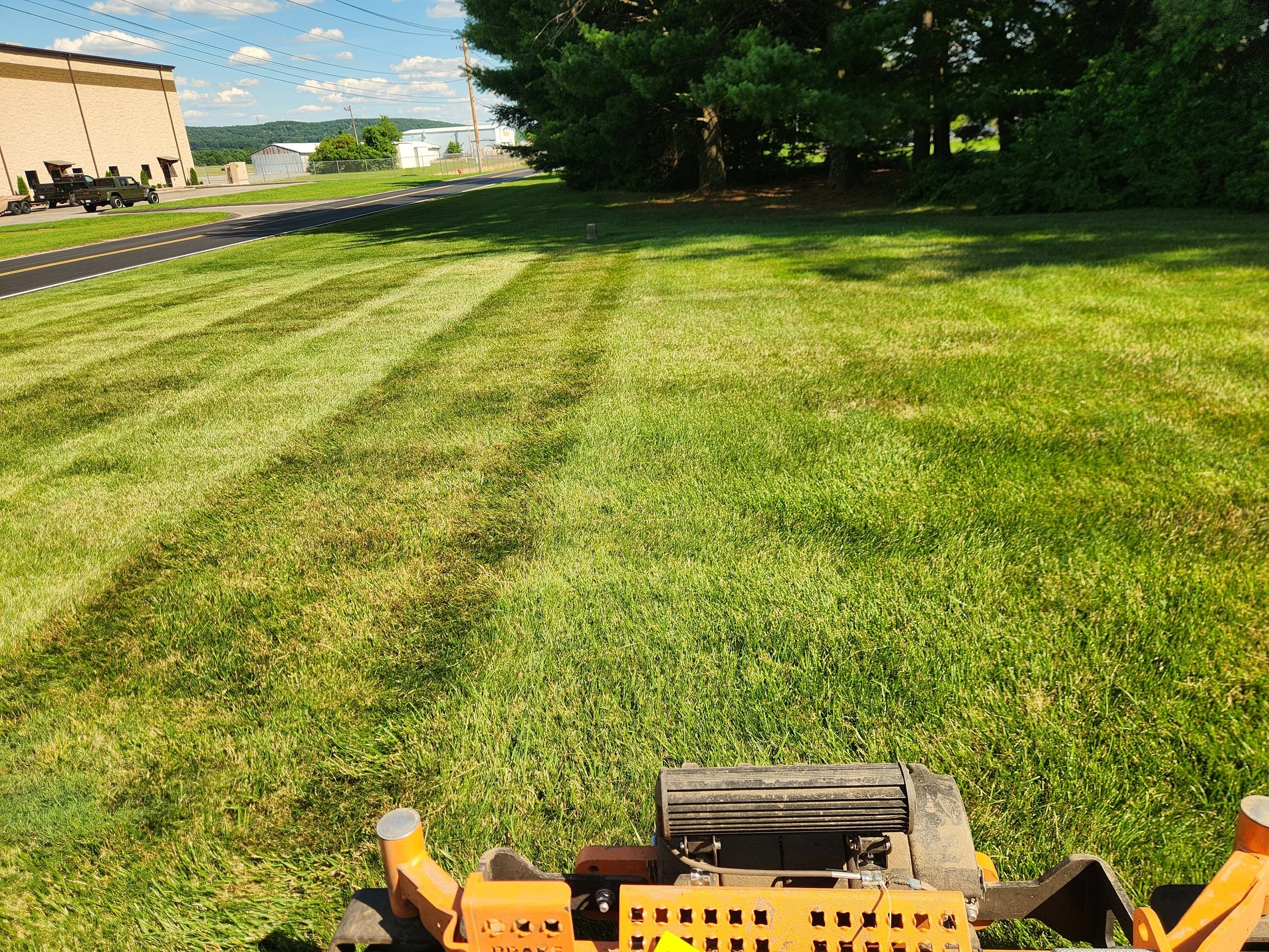 A lawn being mowed with a riding mower on a bright, sunny day with trees and industrial buildings in the background.