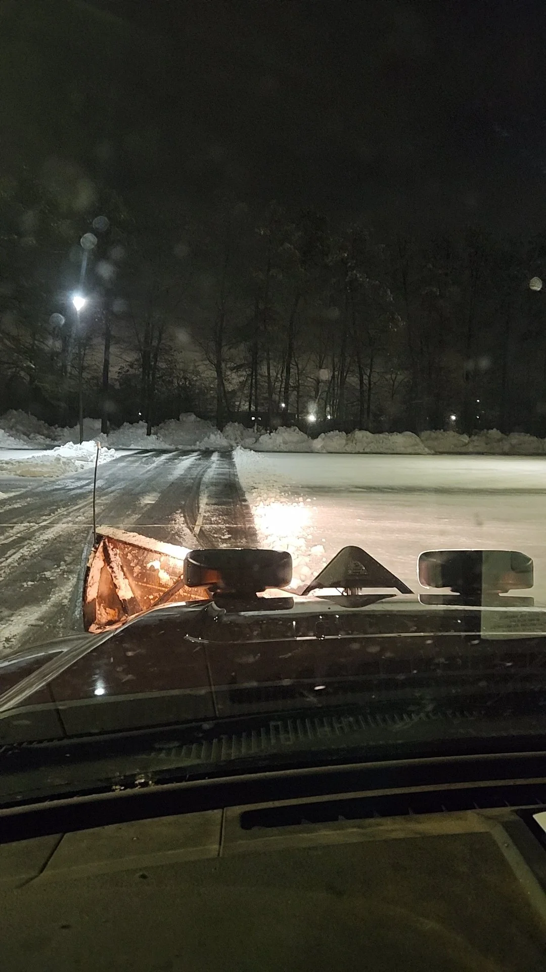 Nighttime view from inside a vehicle with snow on the ground, a snow plow attached to the vehicle's front, and snow-covered trees and streetlights in the background.