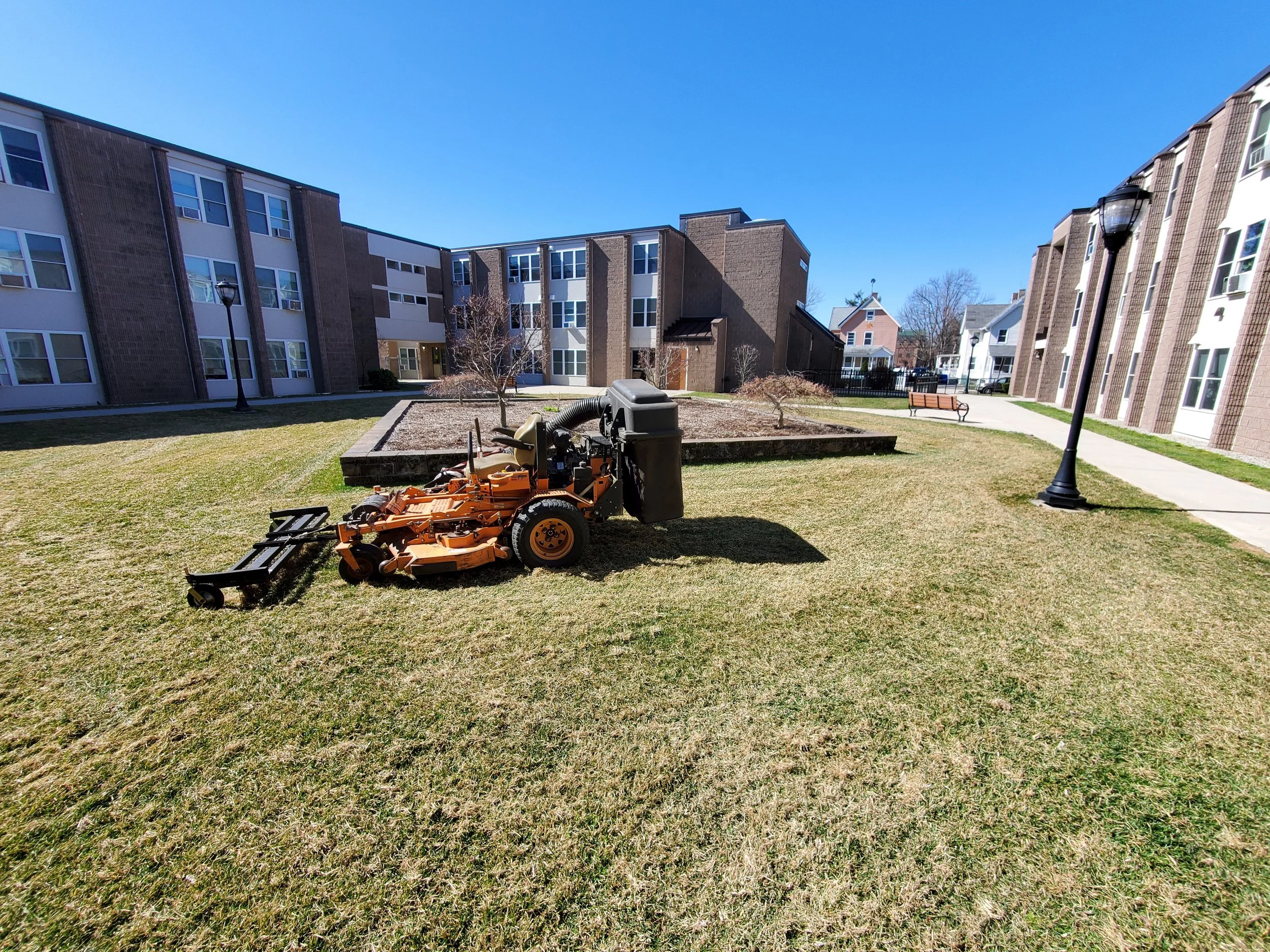 A lawn mower parked on a grassy area in an apartment complex courtyard with pathways, benches, and buildings in the background under a clear blue sky.