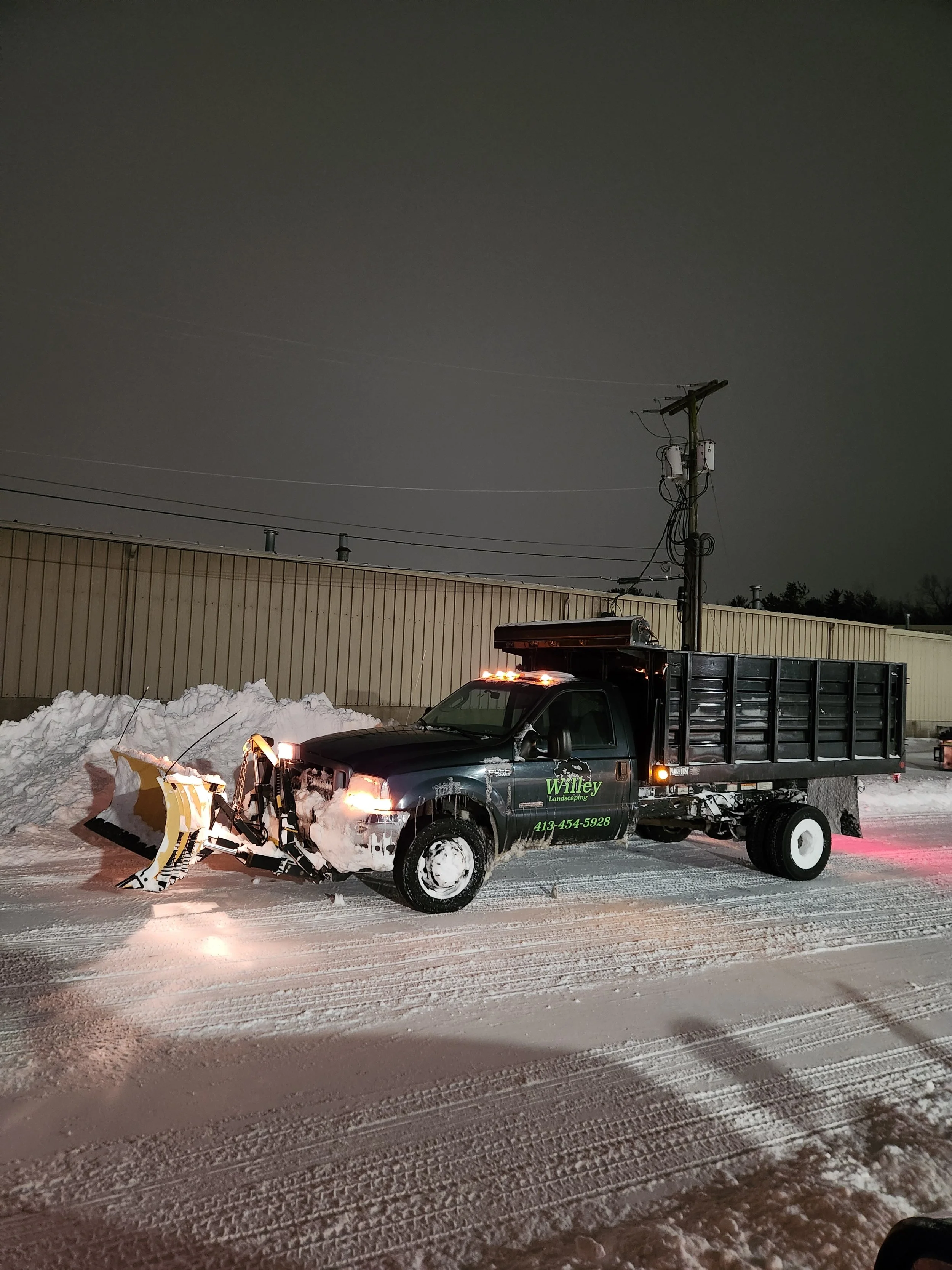 A black landscaping truck with a snowplow attached to the front and a dump bed in the back, parked on a snowy lot at night with a building and utility pole in the background.
