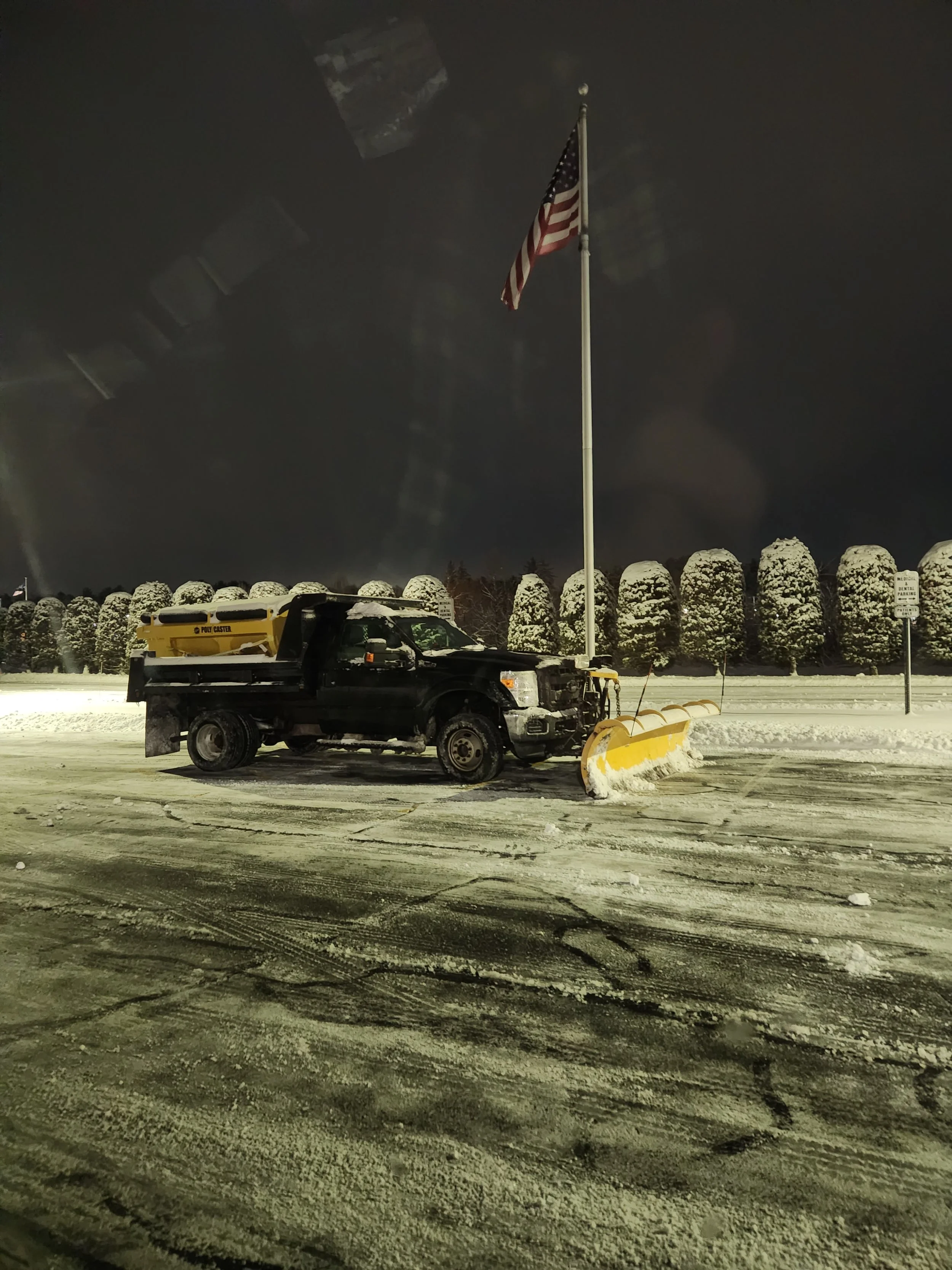 A snow-covered parking lot at night with a snowplow truck and an American flag on a tall flagpole, trees in the background, and a sign on the right side.