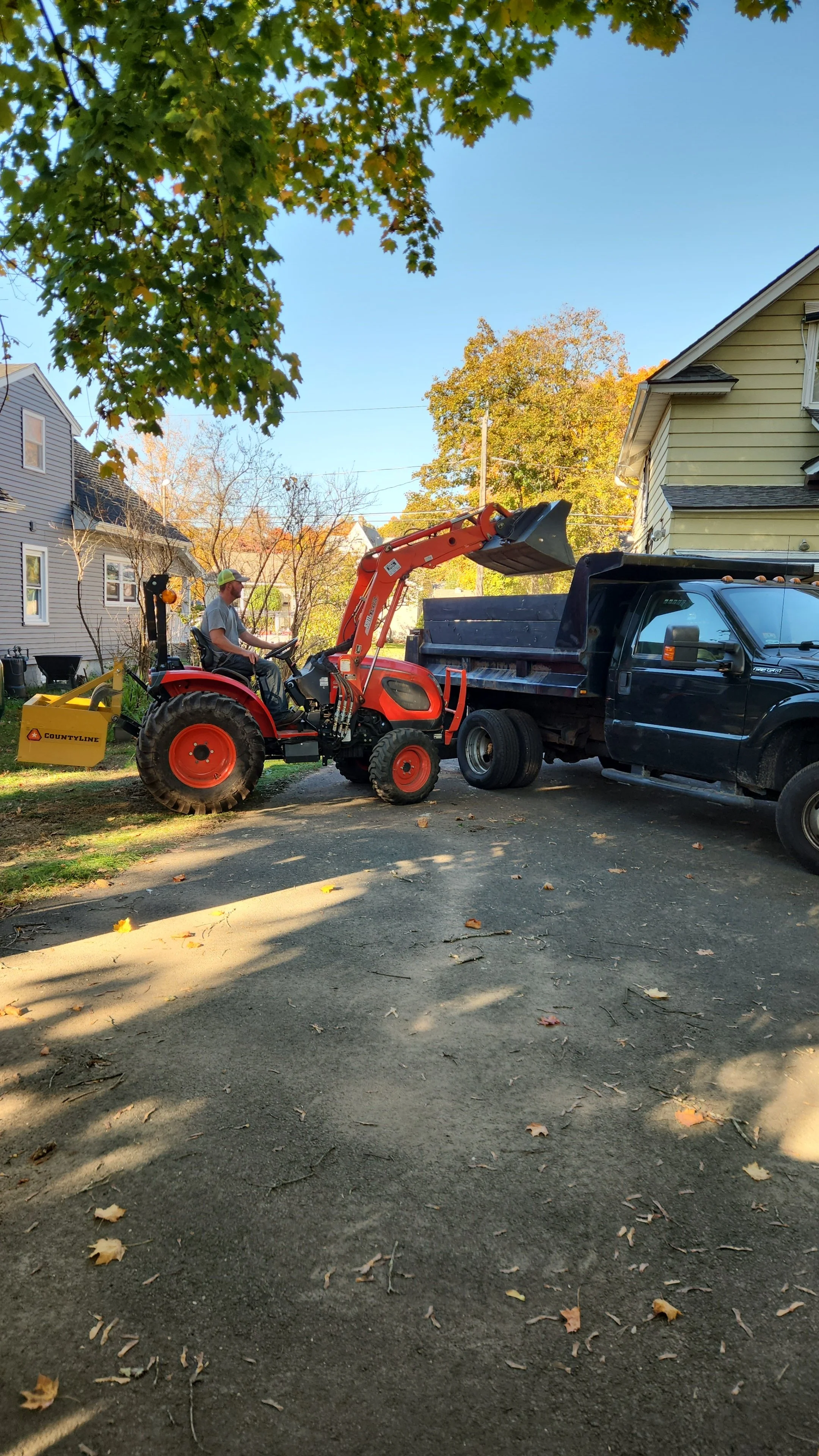 A person operating a red tractor with a front loader attachment, moving a load into a black pickup truck in a residential neighborhood with houses and trees with fall foliage.