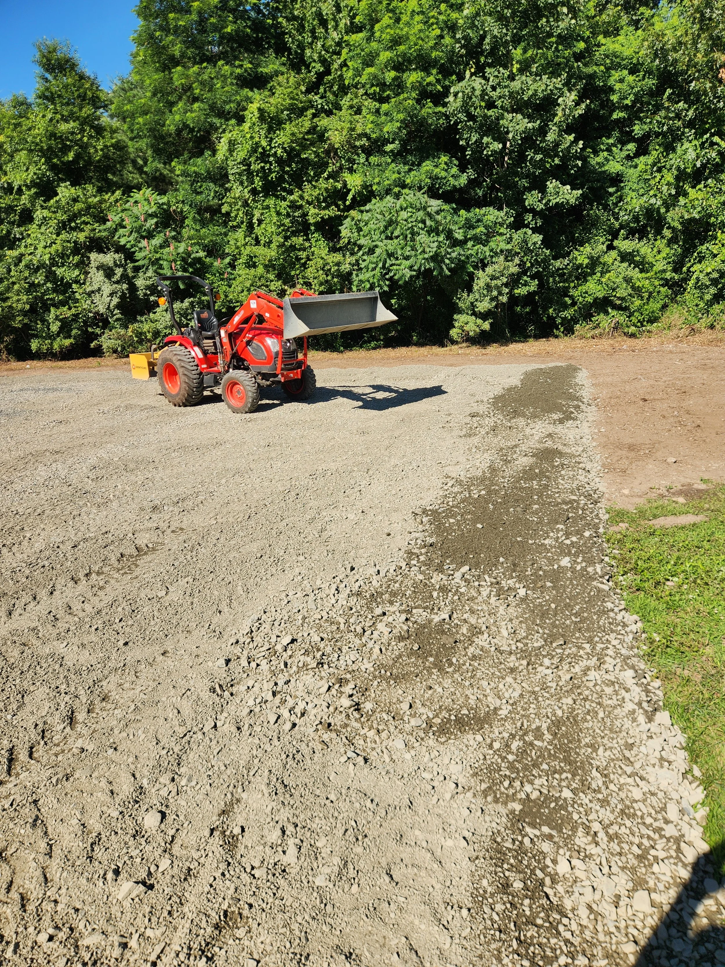 A small red tractor with a front loader attachment parked on a dirt driveway in front of a green, leafy forest.