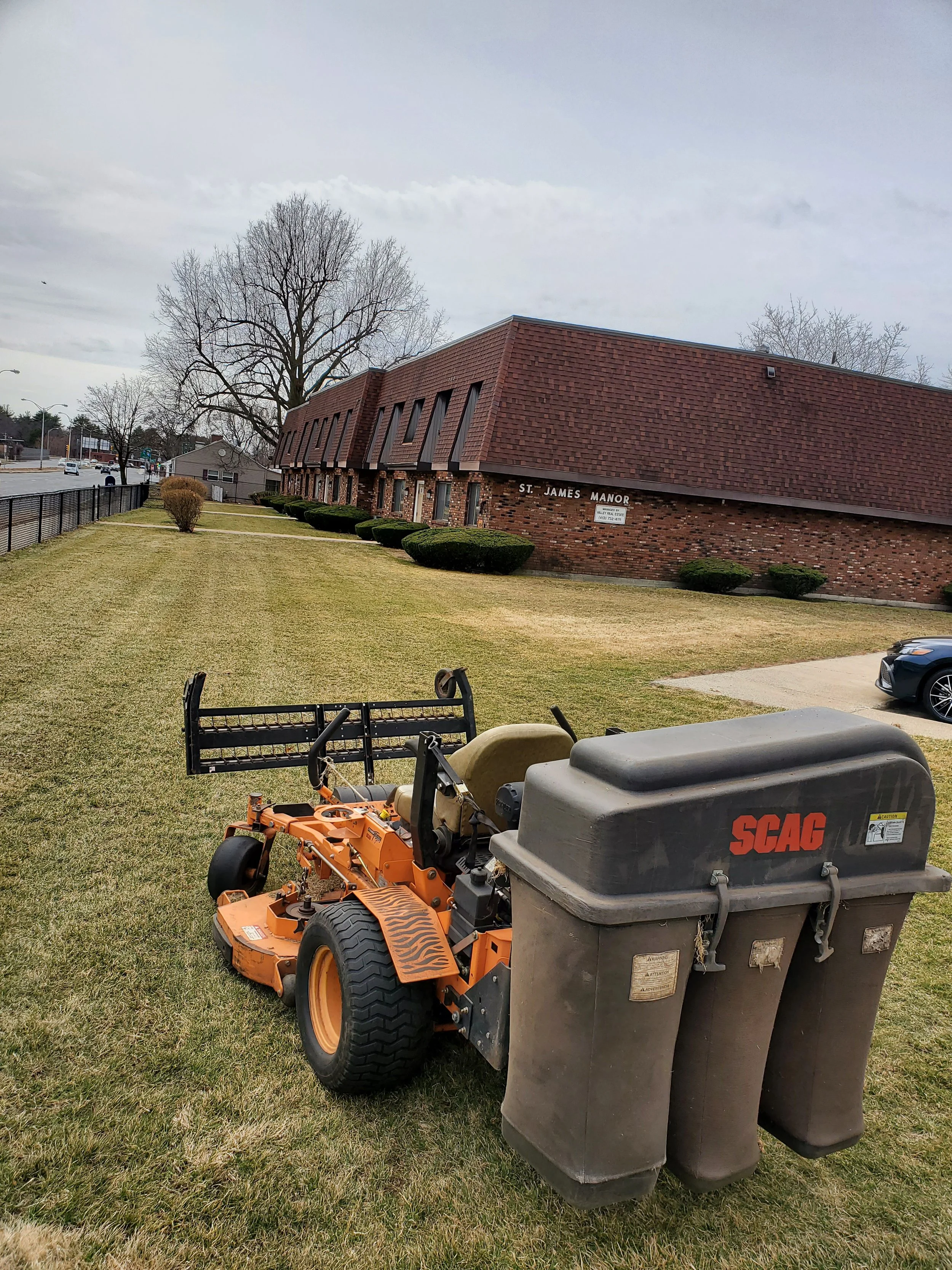 A riding lawn mower with a trash bin attached on a grassy area near a brick building labeled St. James Manor, with some cars parked nearby and trees in the background.