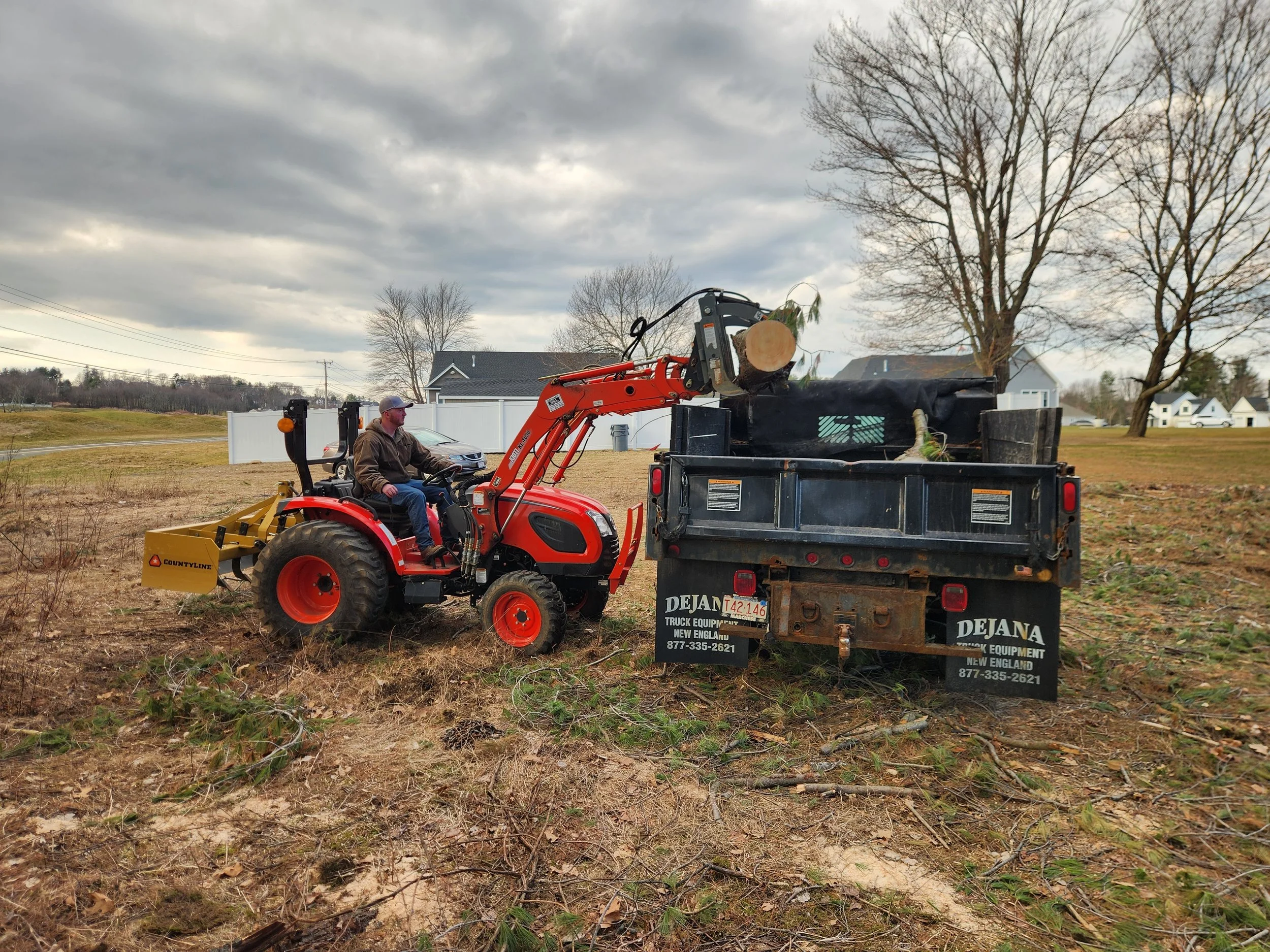 A person is operating a red compact utility tractor with a front loader attachment that is lifting a large log into the bed of a black dump truck on a grassy field. The sky is cloudy, and there are trees and houses in the background.