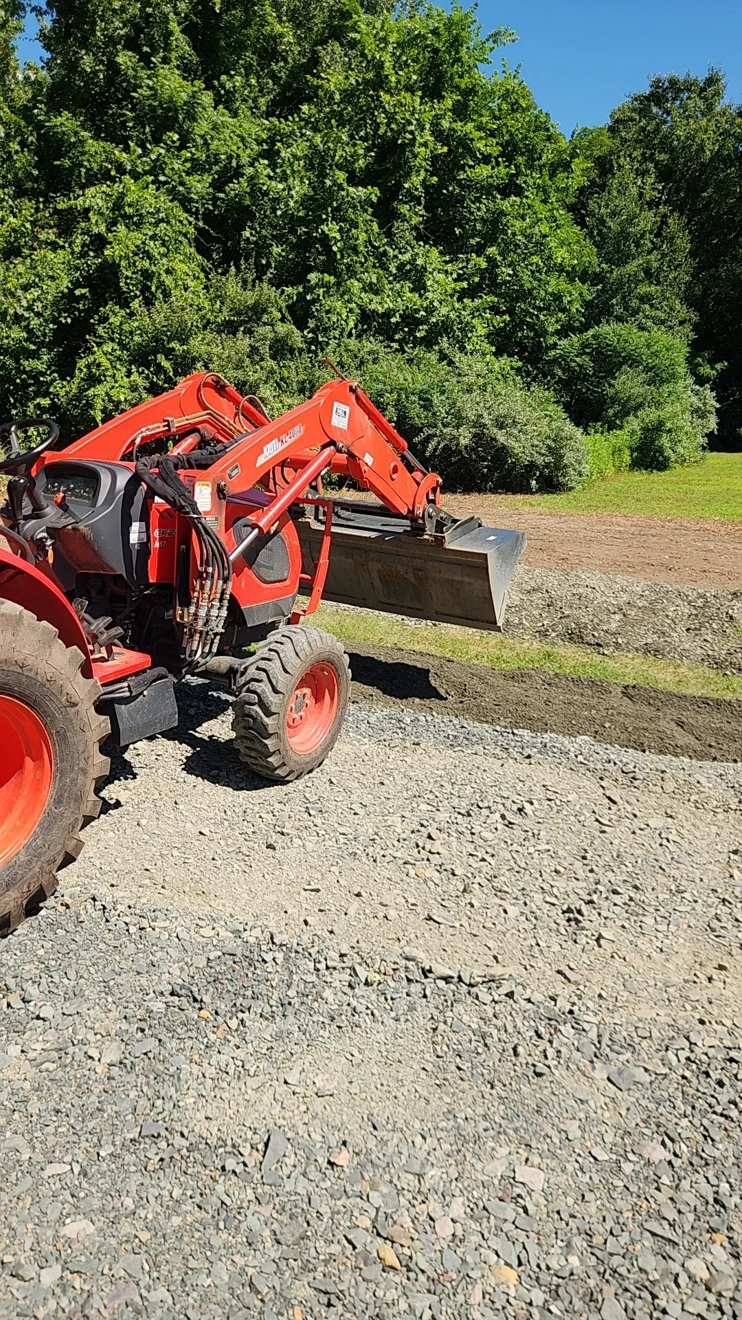 Red compact utility tractor with front loader attachment parked on gravel ground with lush green trees in background.