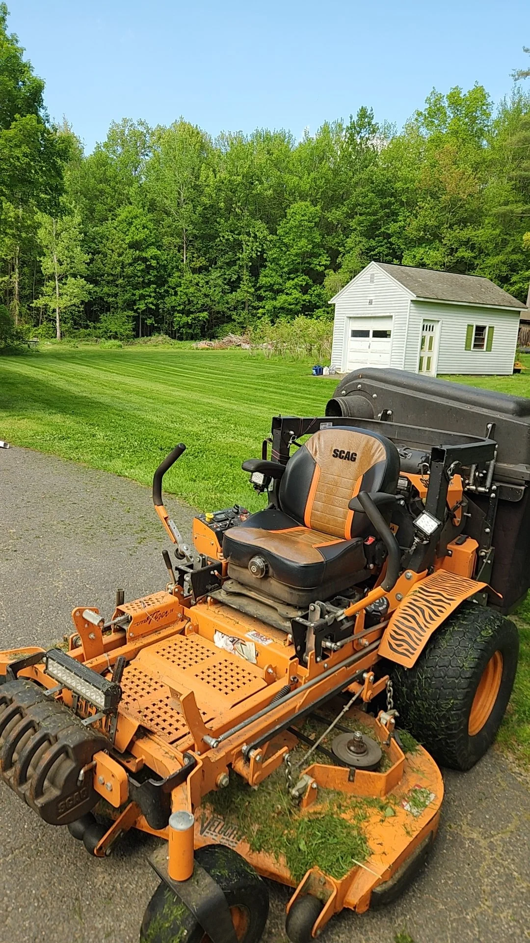 Orange and black lawnmower with a seat and controls, parked on a driveway with grass and trees in the background, and a small white garage building.