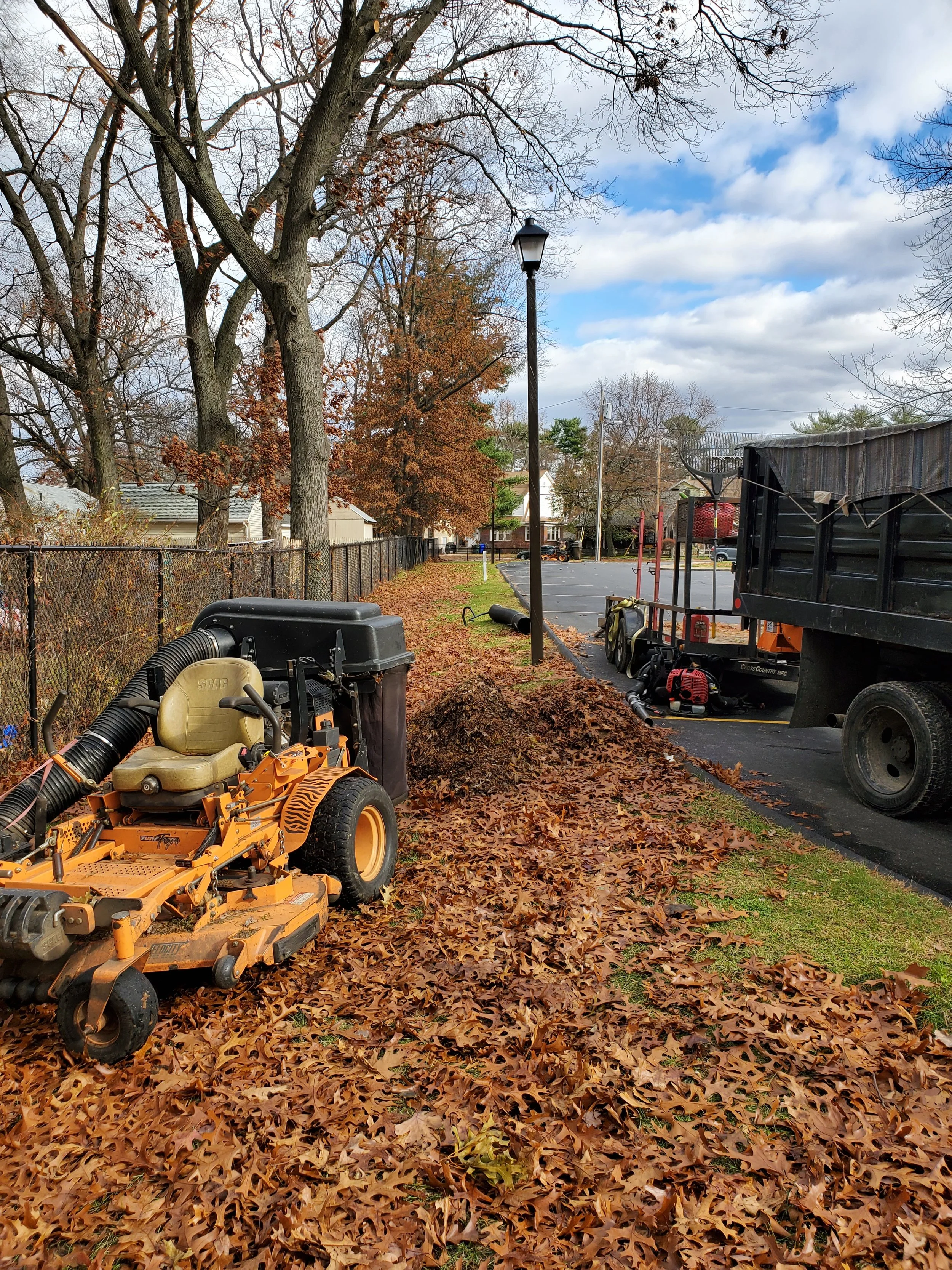 Leaf removal equipment operating along a suburban sidewalk covered with fallen leaves, with trees, a fence, and a parking lot in the background.