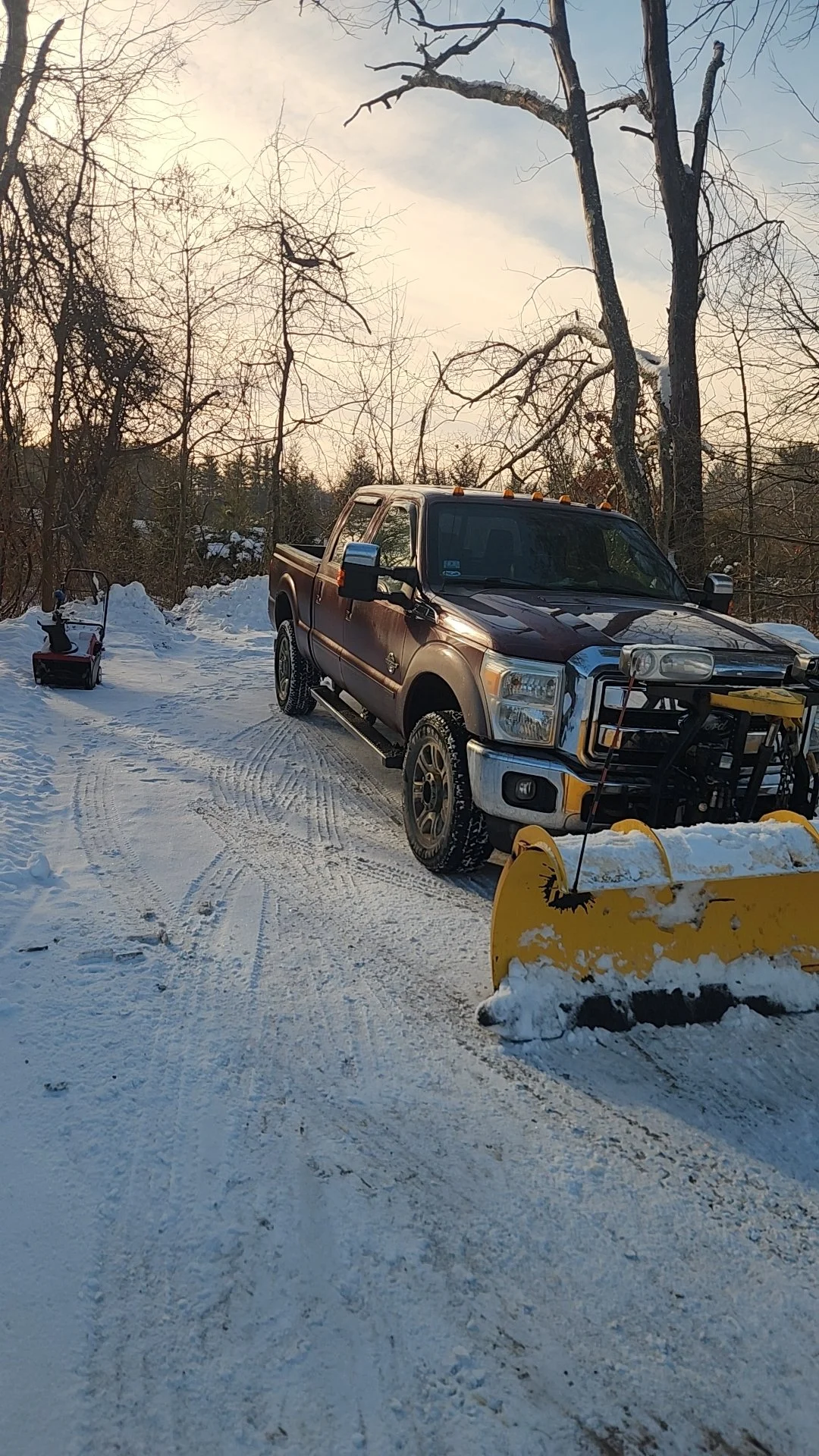 A pickup truck equipped with a snow plow attachment on a snowy country road. A snow blower is parked nearby. Bare trees are visible, suggesting winter.