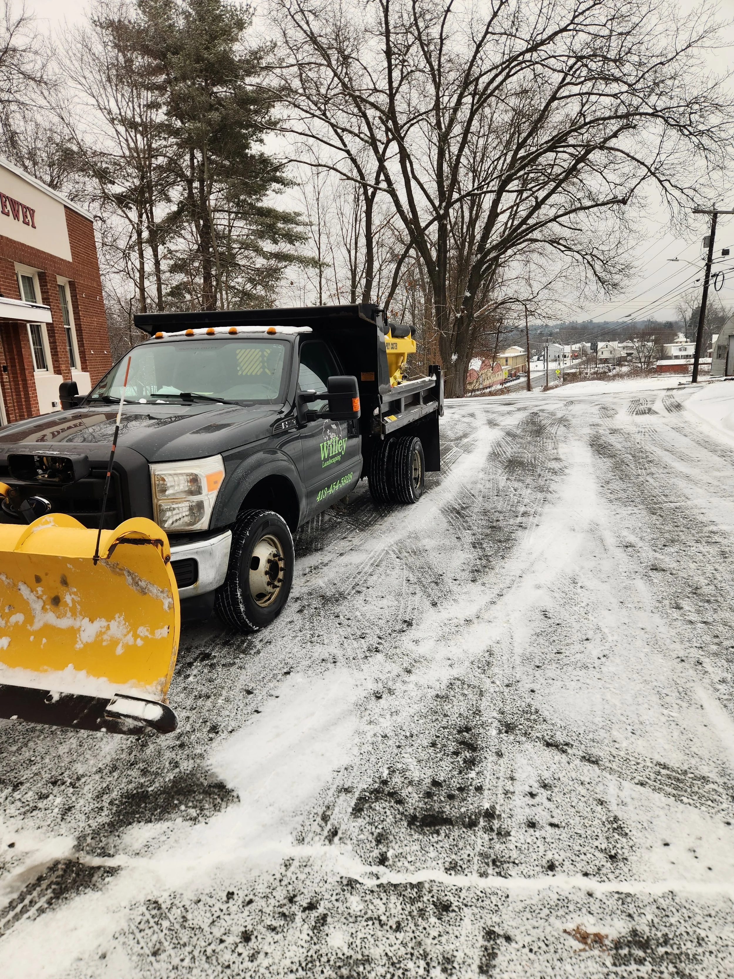 A black snow plow truck with yellow equipment in back, parked on a snowy street near a brick building under leafless trees.