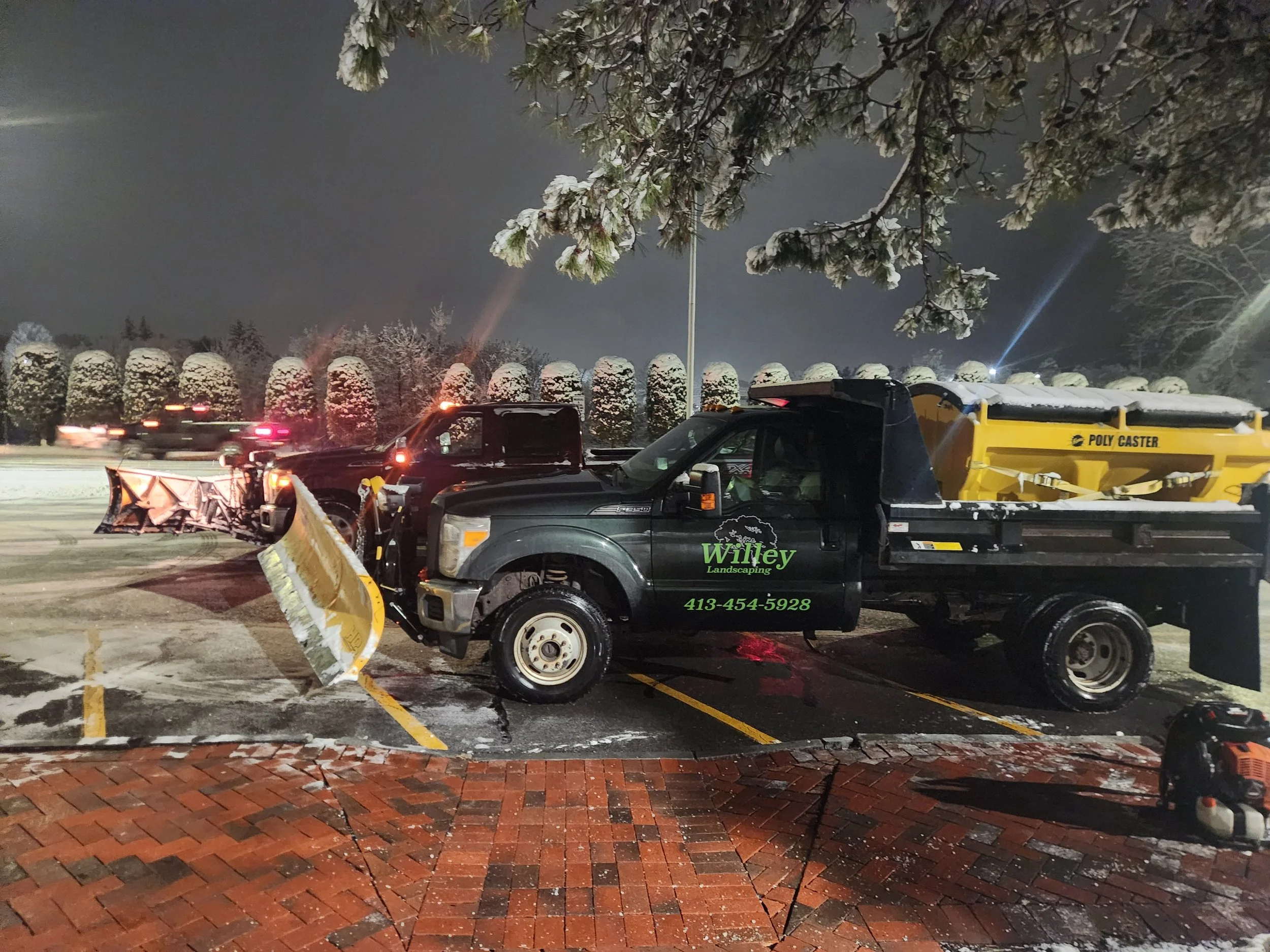 A snow-covered parking lot at night with a landscaping truck and snow plow blade attached, clearing snow. Snow-covered trees are visible in the background.