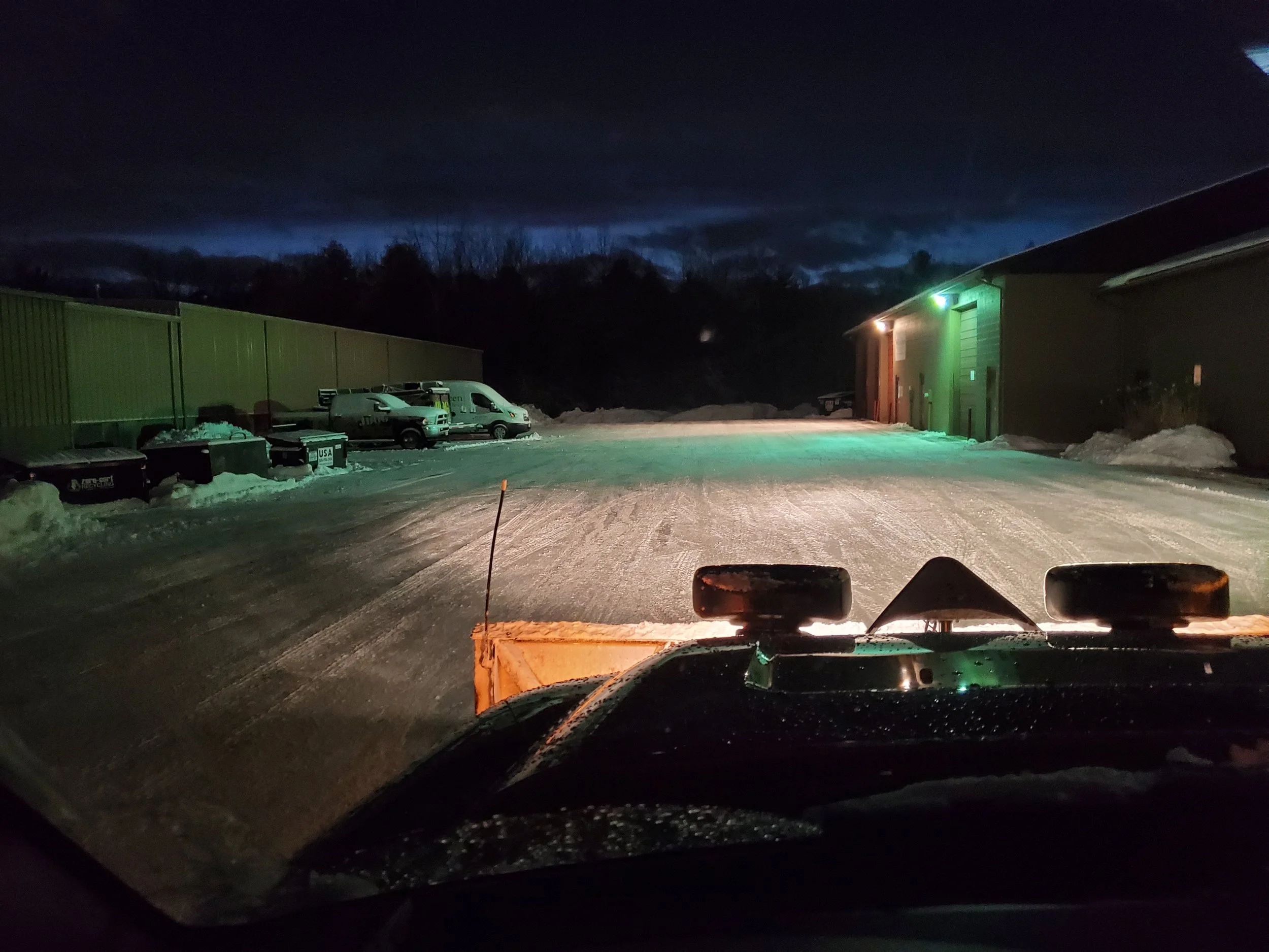 Nighttime view from vehicle showing a snow-covered parking lot with several parked vehicles, a green building with colorful lights, and dark sky with some clouds.