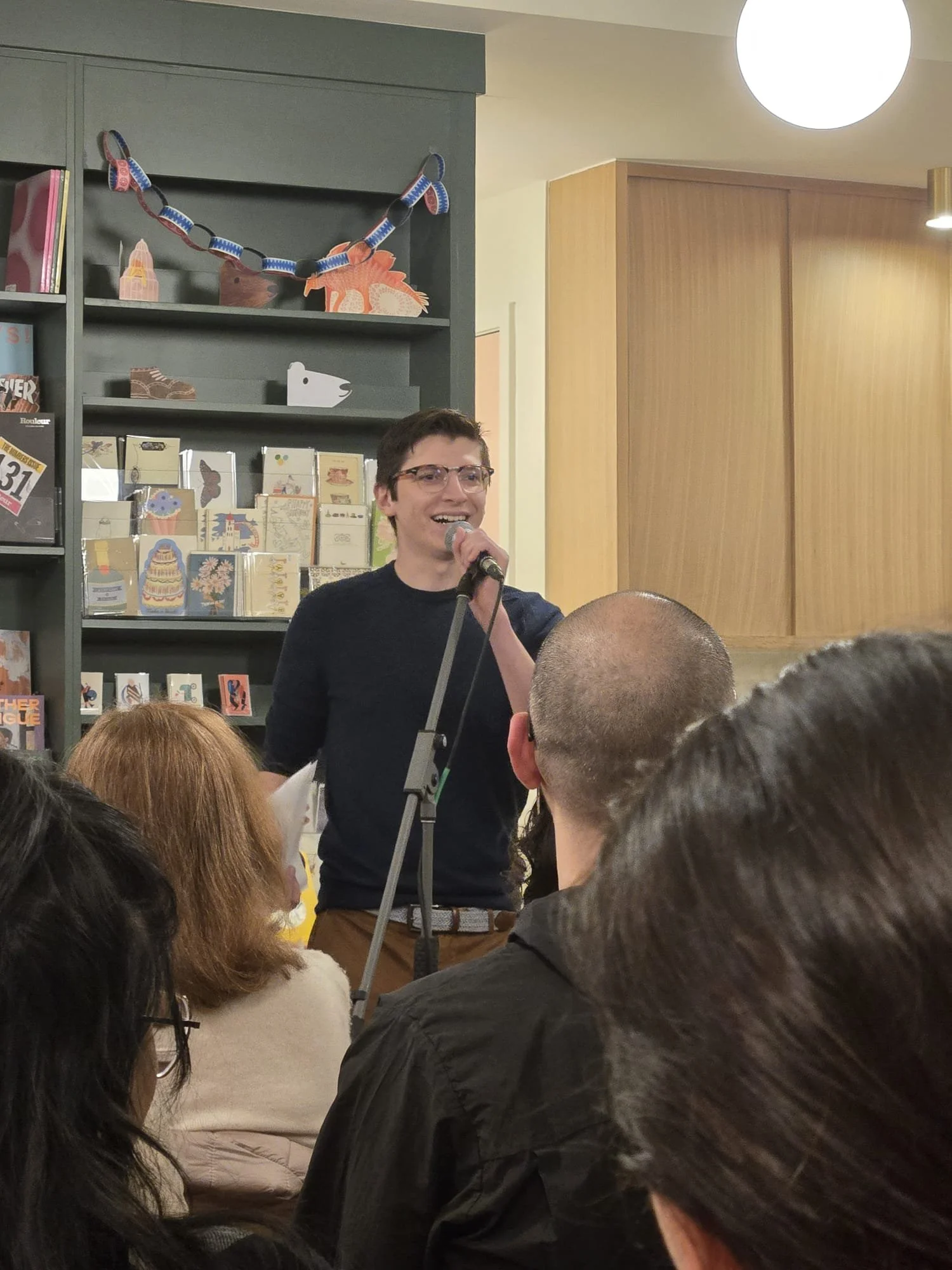 A man with glasses and dark hair speaking into a microphone at a gathering, with an audience seated in front of him, in a room decorated with books and crafts on shelves and a paper chain garland.