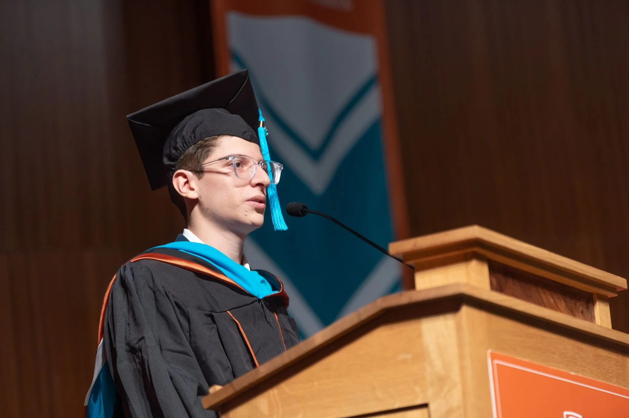 A young man in graduation regalia, including a black cap and gown with a blue sash, stands at a podium speaking into a microphone during a graduation ceremony.