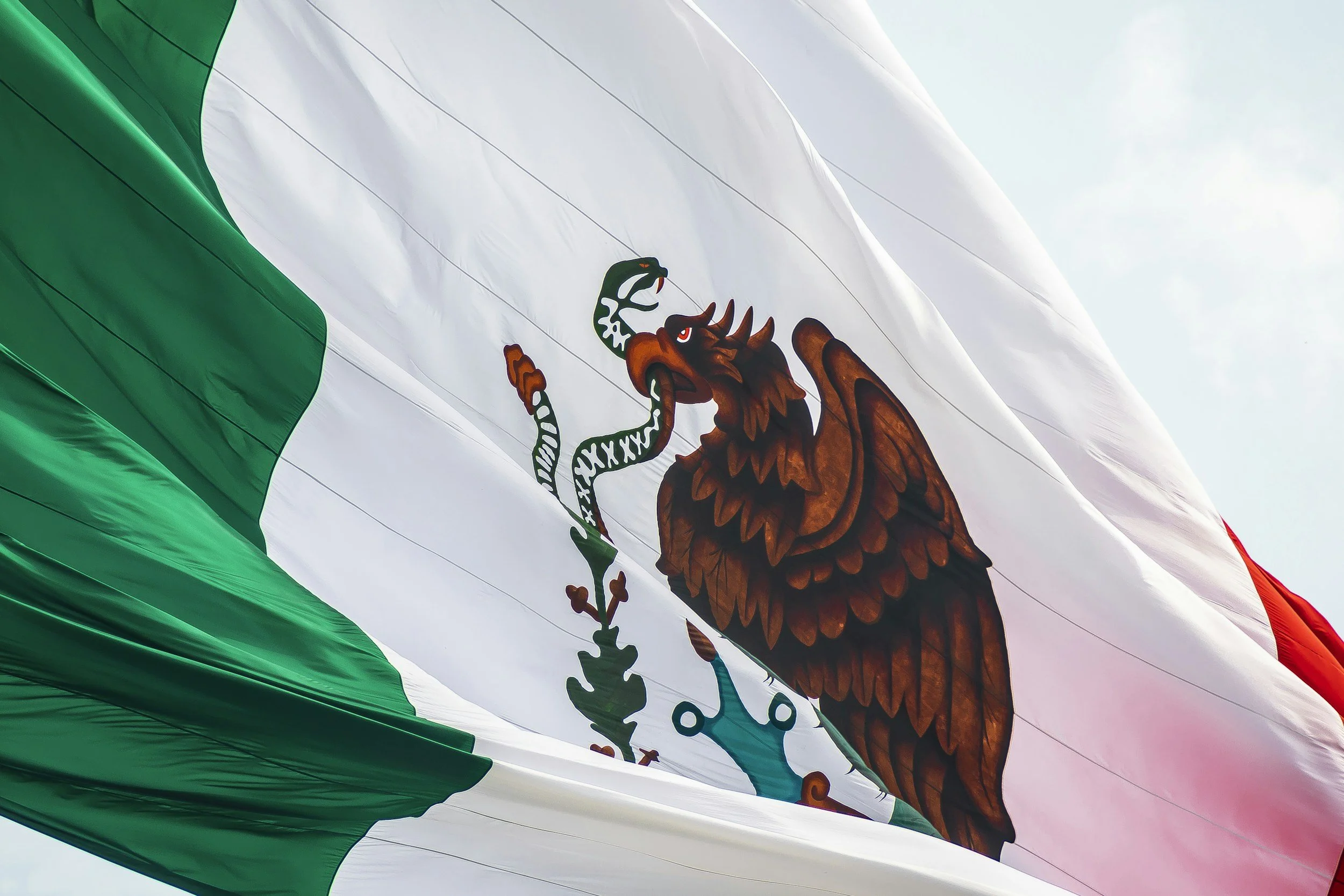 Close-up of the Mexican flag, featuring a green, white, and red vertical stripe with the national coat of arms in the center, displaying an eagle holding a snake in its beak and talon, perched on a cactus.