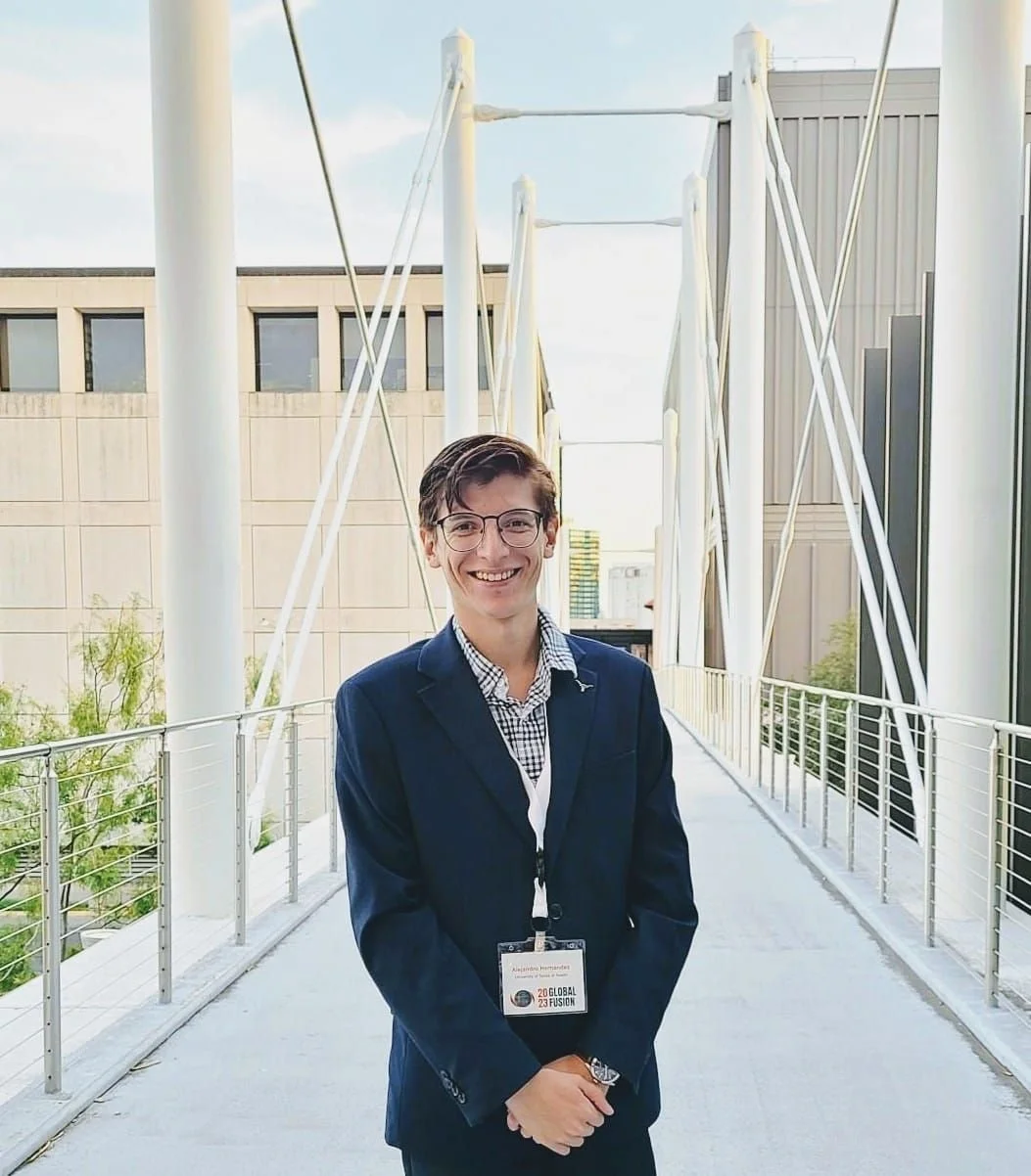 A young man wearing glasses, a navy blazer, and a lanyard with a badge standing on a modern pedestrian bridge with steel cable supports and city buildings in the background.