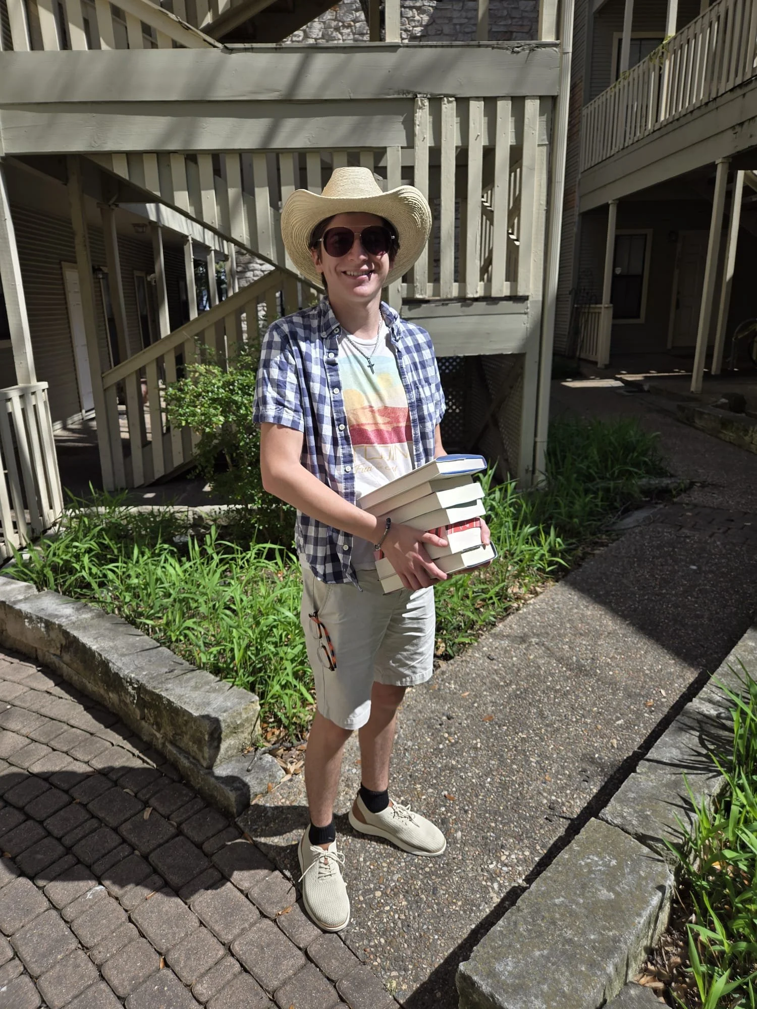 A young man standing outside on a sidewalk, smiling, wearing a straw hat, sunglasses, a checkered shirt over a graphic T-shirt, beige shorts, white shoes, and black socks, holding a stack of books.