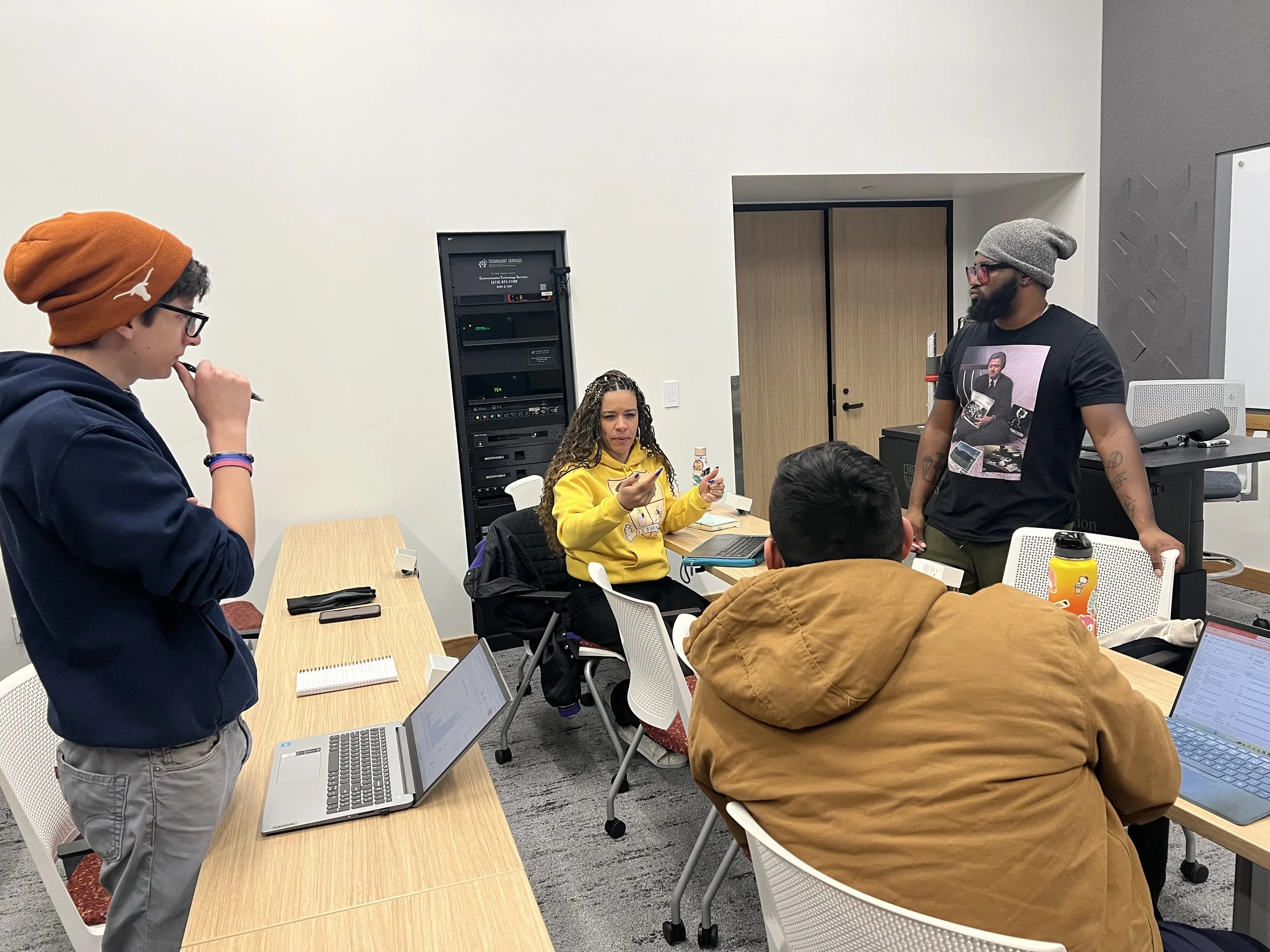 People in a meeting room engaged in a discussion. One woman in a yellow hoodie holds a phone, while a man in a gray beanie and black T-shirt stands. Two seated individuals have laptops open, and another young man in a brown jacket is sitting with his back to the camera.