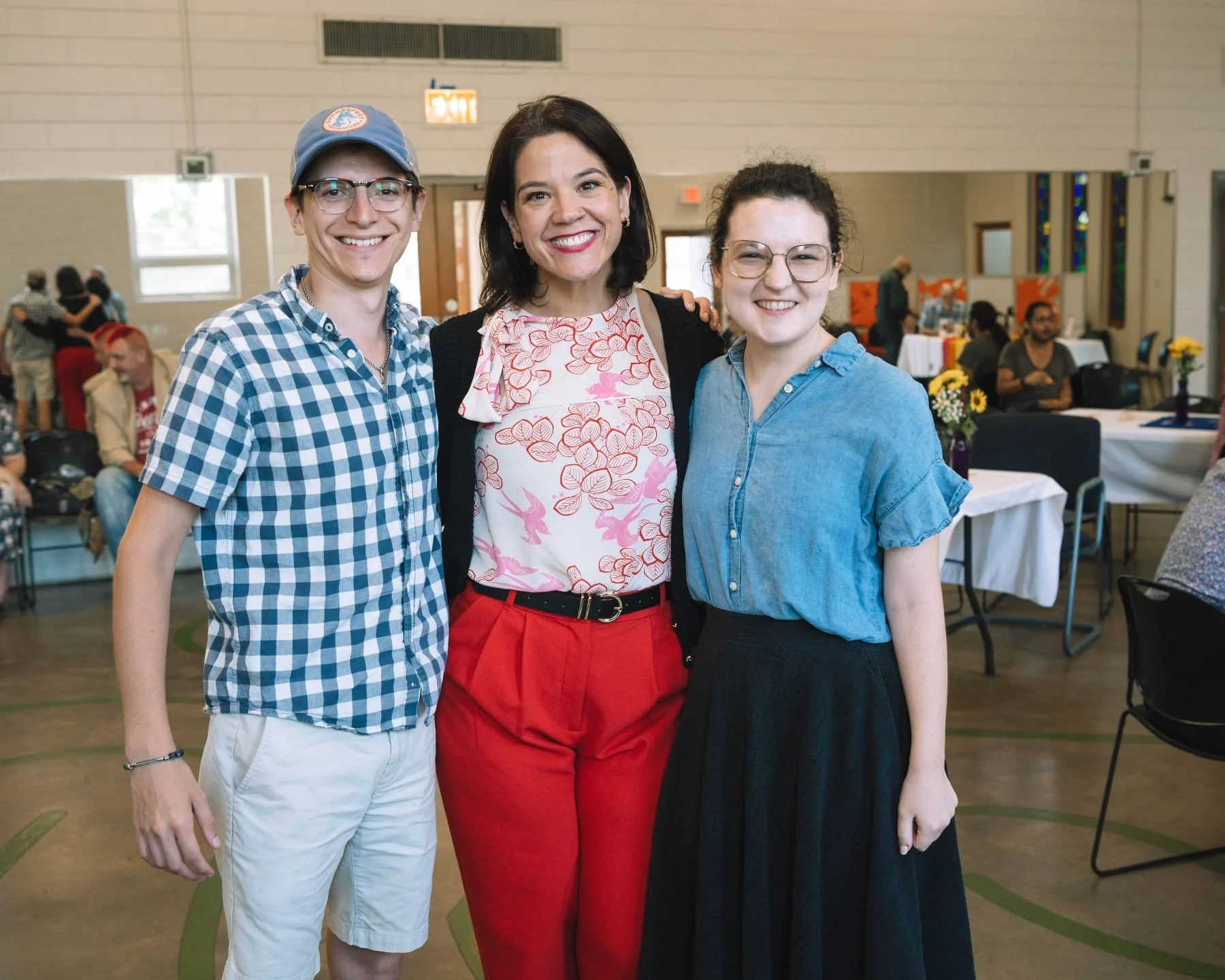 Three women smiling and standing together in a community center or event hall, with tables and people in the background.