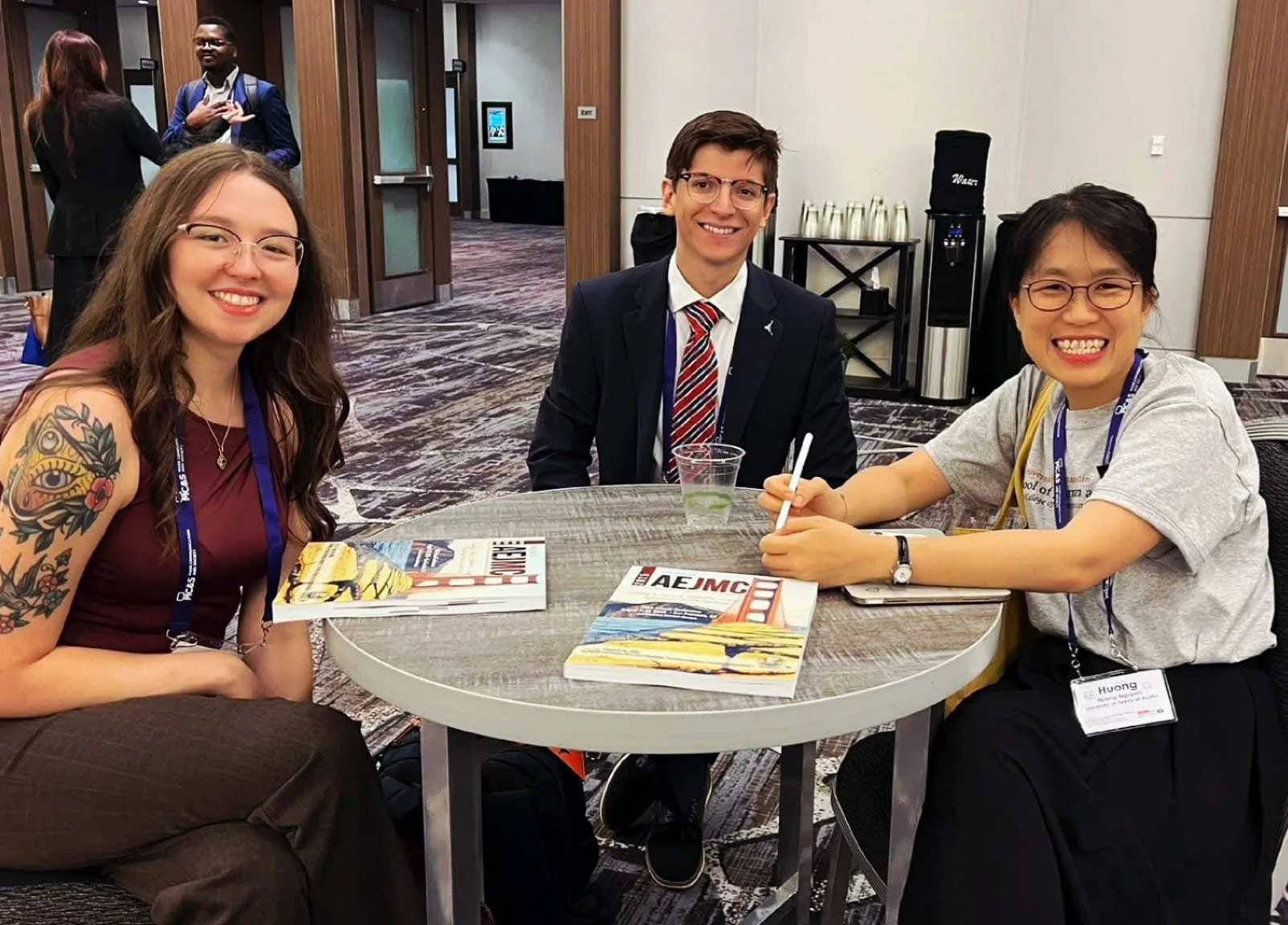 Three people sitting at a round table in a conference room smiling at the camera. The table has magazines and a plastic cup. Behind them are two women talking, and the background shows conference room doors and a water cooler.