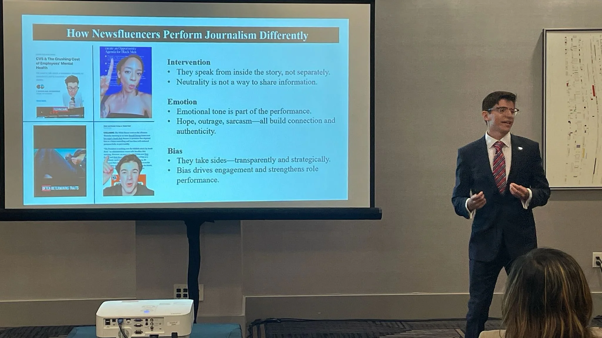 A man in a suit and tie giving a presentation in front of a large screen in a conference room, with a woman in the foreground listening.