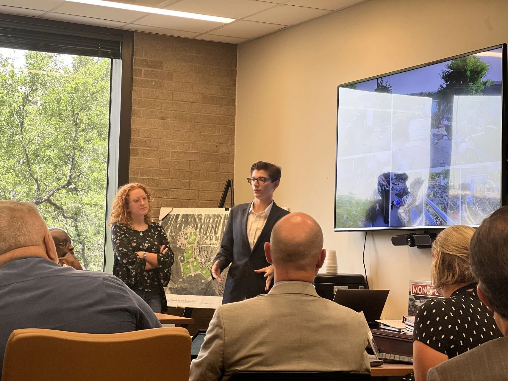 A man in a dark suit and glasses is giving a presentation in a conference room. Several people are seated and listening, with a woman standing near a large map or plan. A large window shows green trees outside, and a big screen displays a video or image.