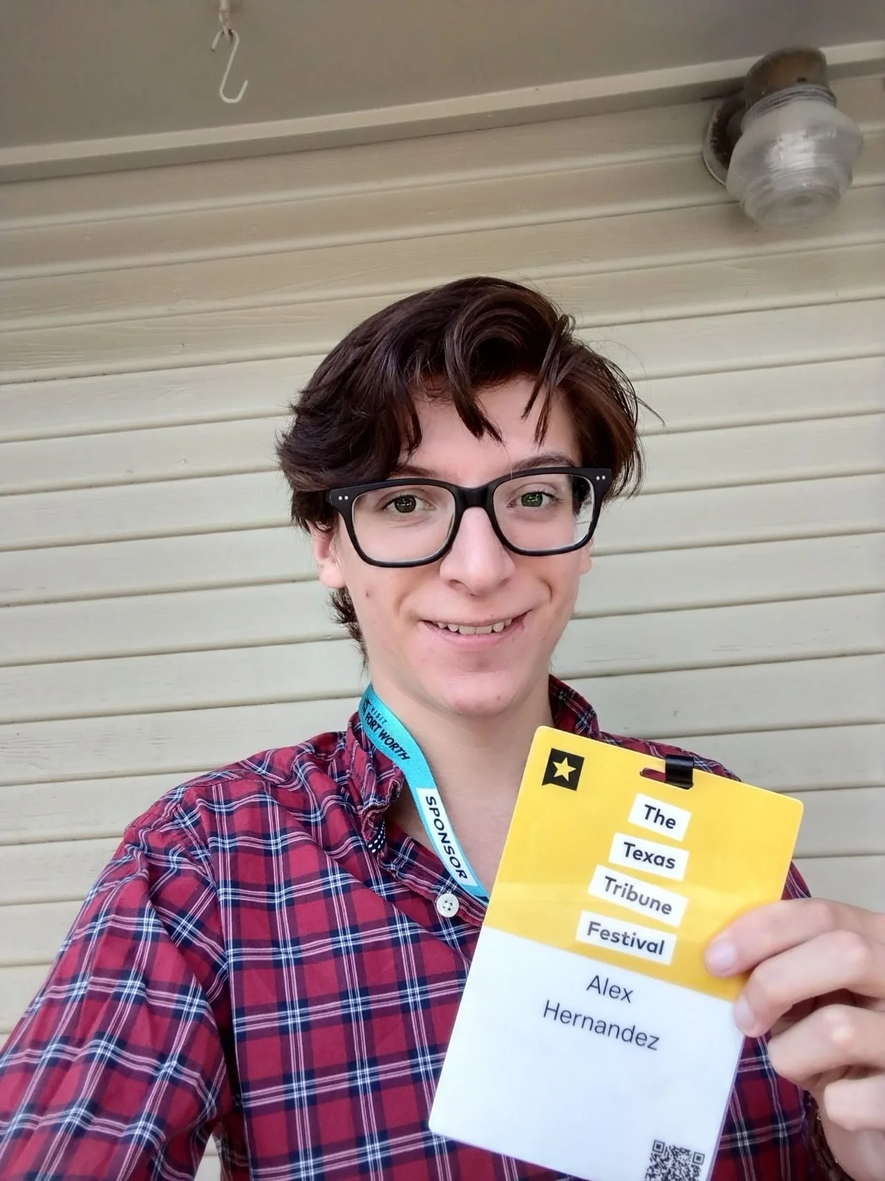 A young man with glasses and brown hair smiling, holding a festival badge with the text "The Texas Tribune Festival" and his name, Alex Hernandez, written on it.