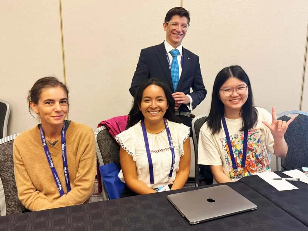 Four people posing for a photo, three women sitting at a table and one man standing behind them, all smiling at a conference or meeting.