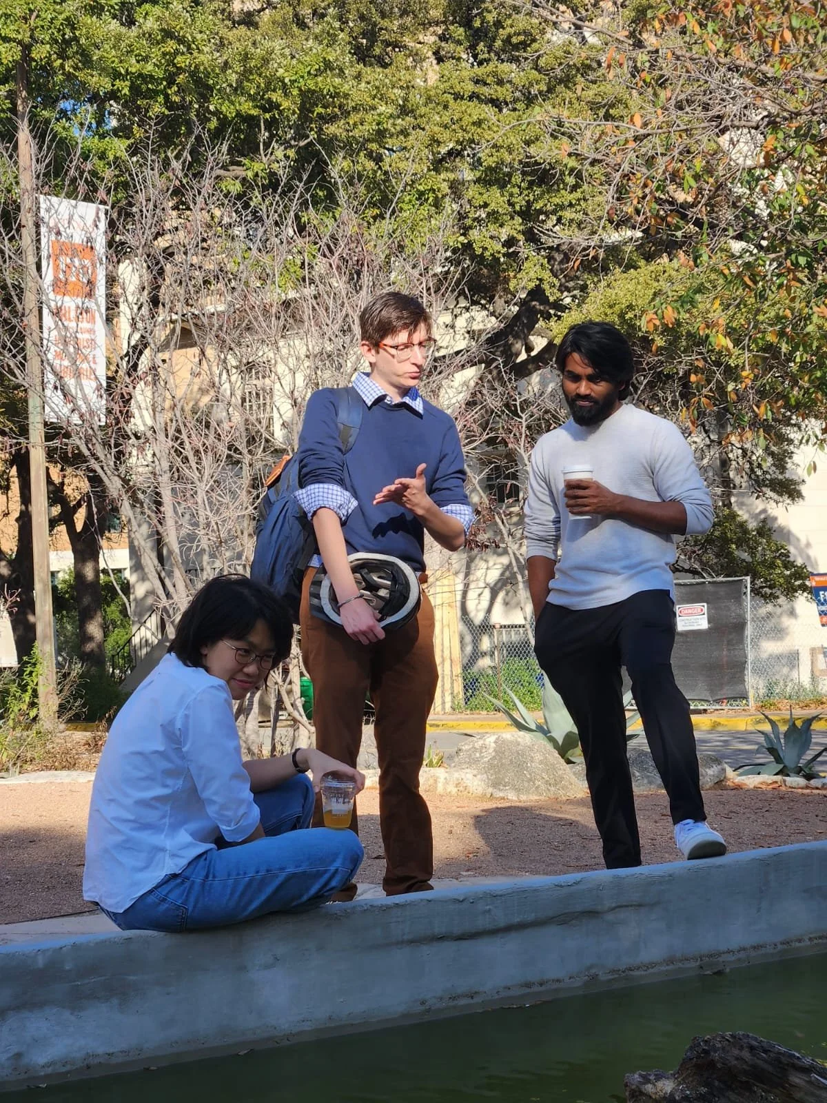 Three people standing outdoors near a pond, two men talking and a woman sitting on the ground holding a drink, in a park with trees and a chain-link fence in the background.