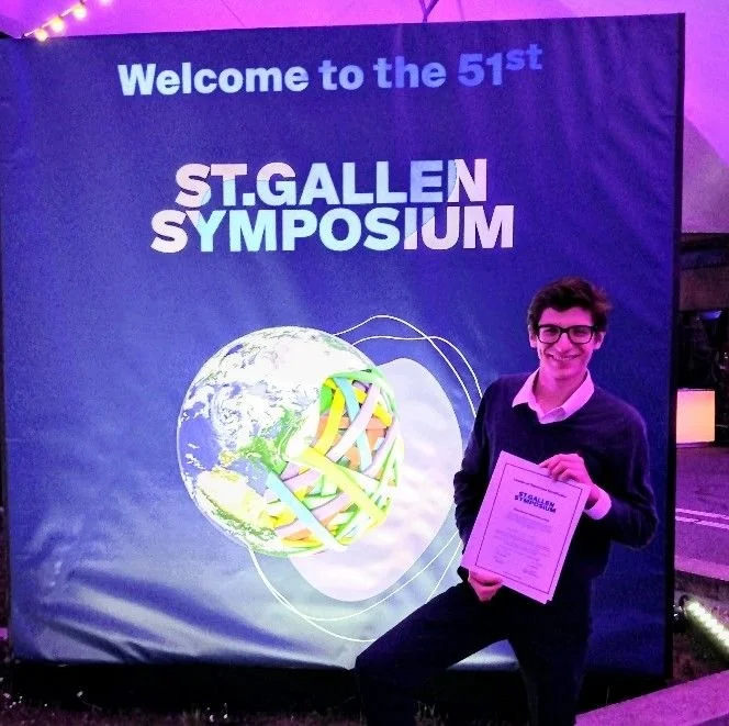 A young man in glasses and a dark sweater, holding a certificate, standing next to a banner that reads "Welcome to the 51st St. Gallen Symposium" with an image of the Earth surrounded by colorful ribbons.