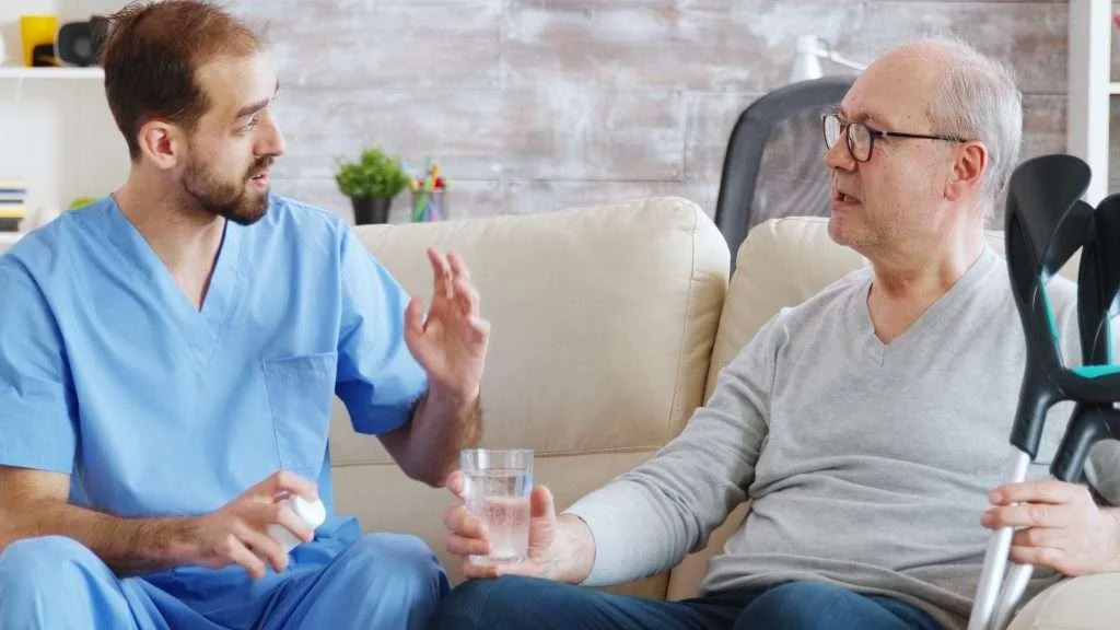 A young male nurse or caregiver with a beard and blue scrubs talking to an older man with gray hair, glasses, and a gray shirt, sitting on a beige couch in a bright, modern living room. The nurse is gesturing with his hand while holding a pill bottle, and the older man is holding a glass of water.