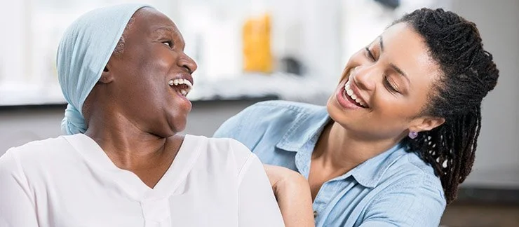 A woman in a medical gown and head covering laughing with a caregiver in a hospital or clinic setting.