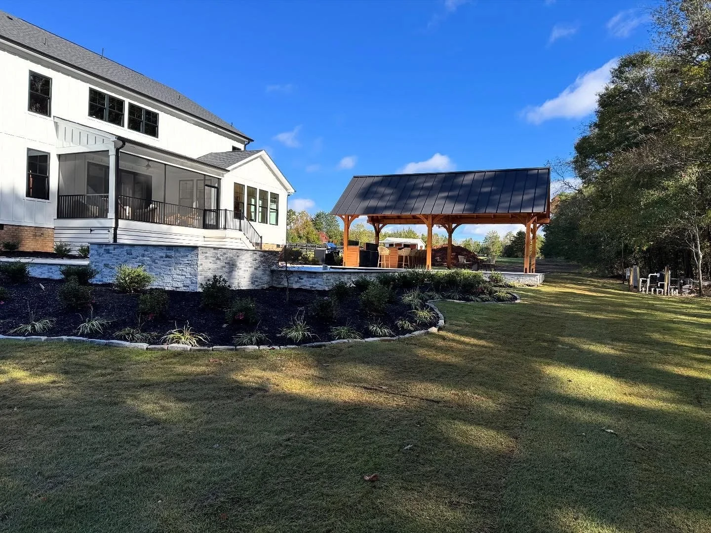 Yard redesign with stone patio, mulched garden beds with ornamental plants installed, a stone paver border, and fresh sod installation behind white house with a pavilion 