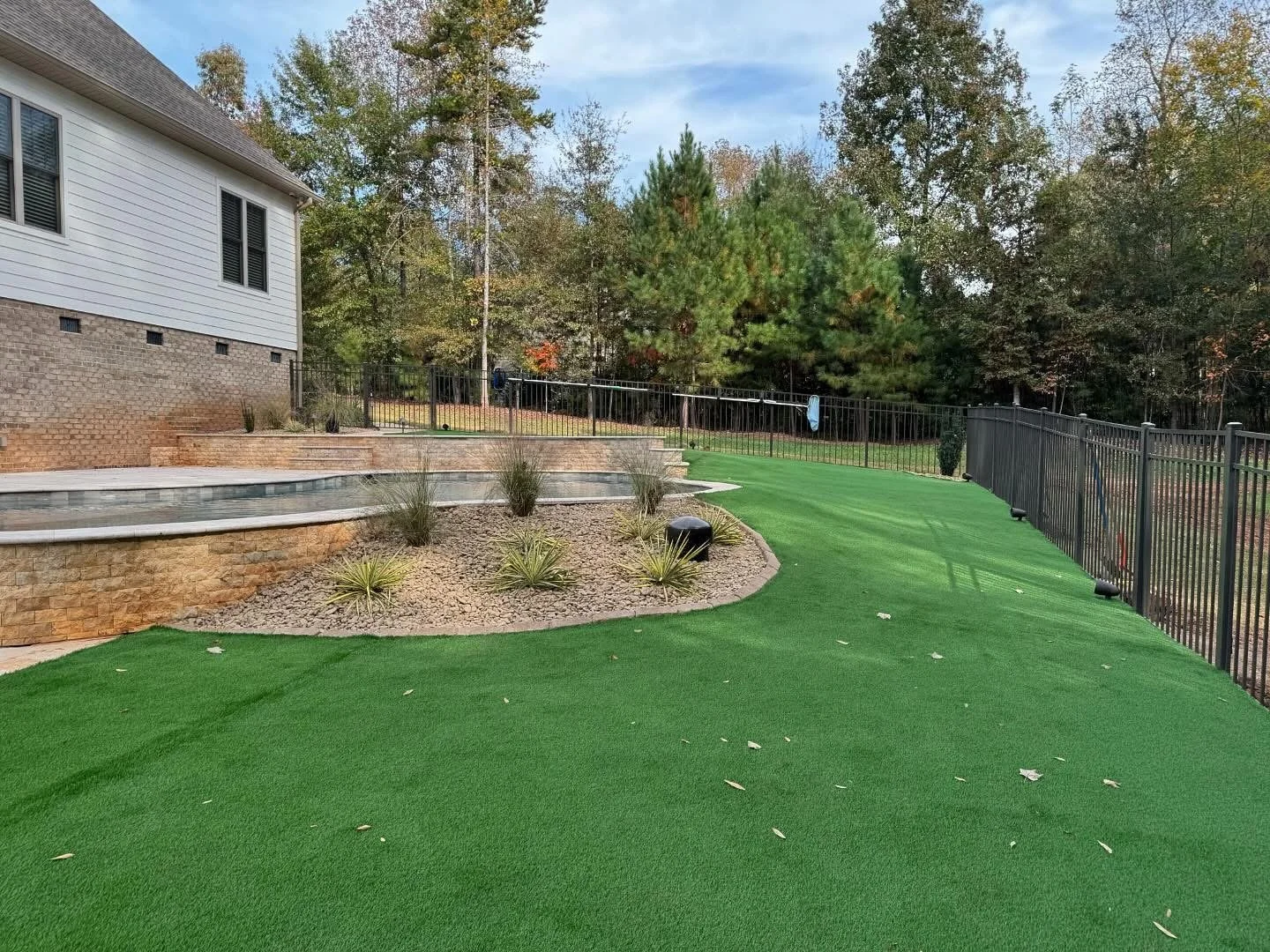 Artificial turf installation and garden bed redesign with river rock and ornamental plants around stone patio 