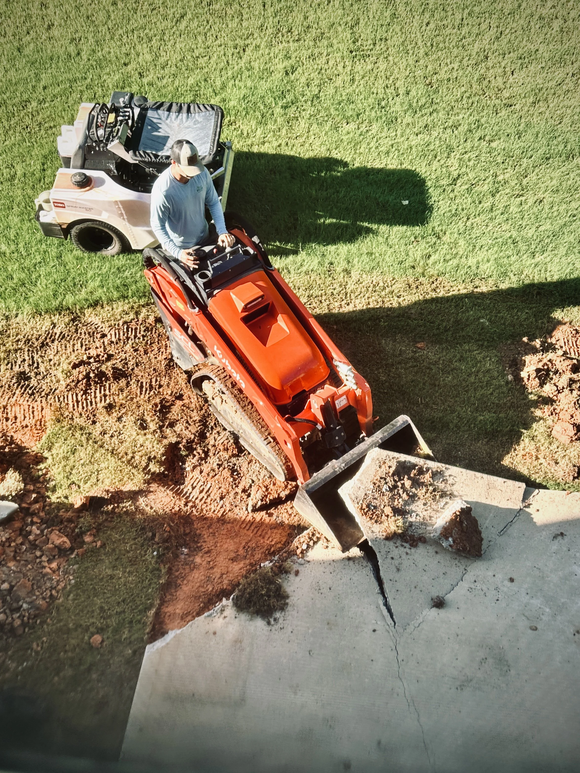 Overhead image of a landscaper on orange soil cultivator grading a lawn with spray master machine in background