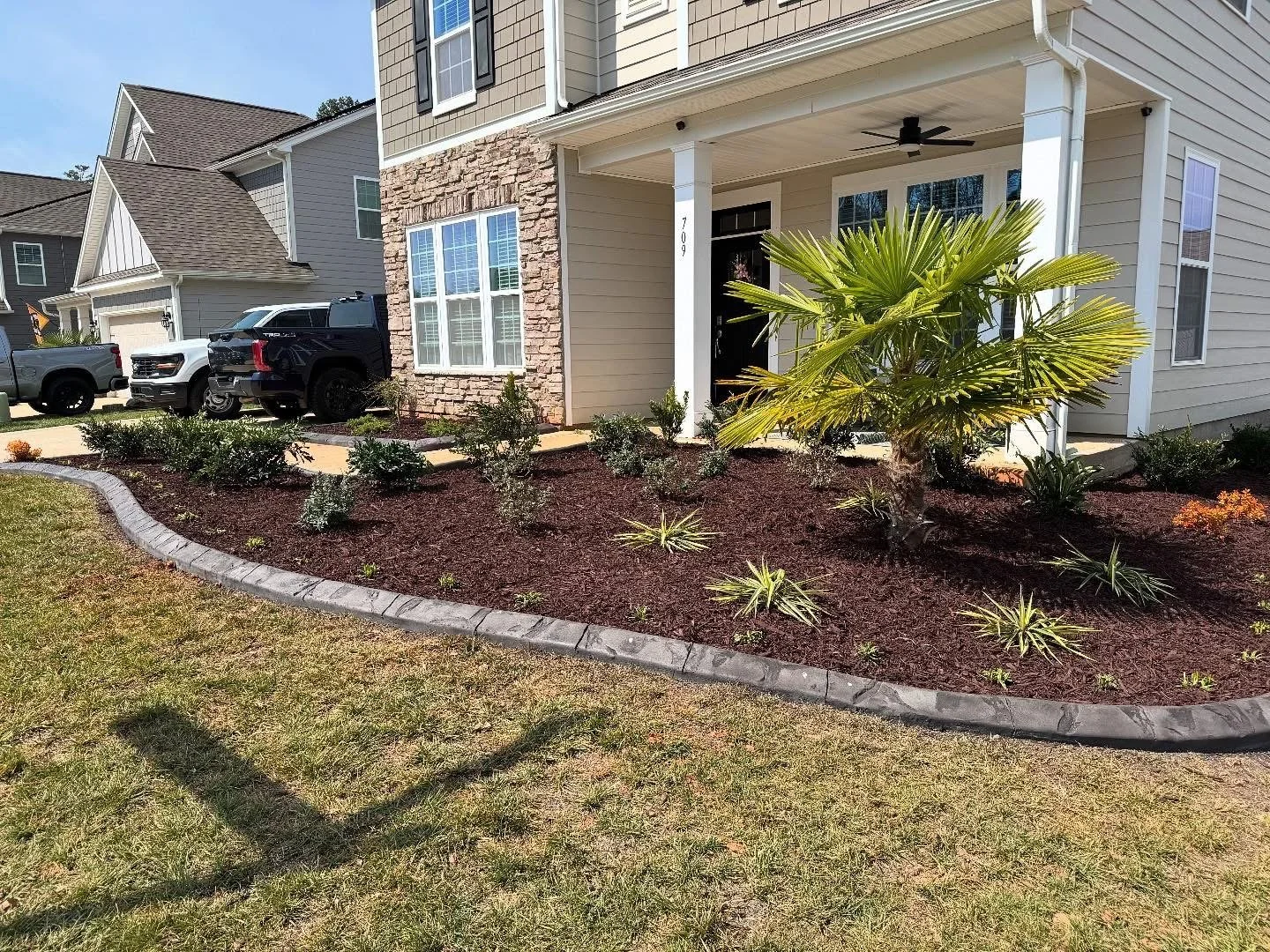 Mulched garden bed in front of home with stone paver border and plants, flowers, and palm tree