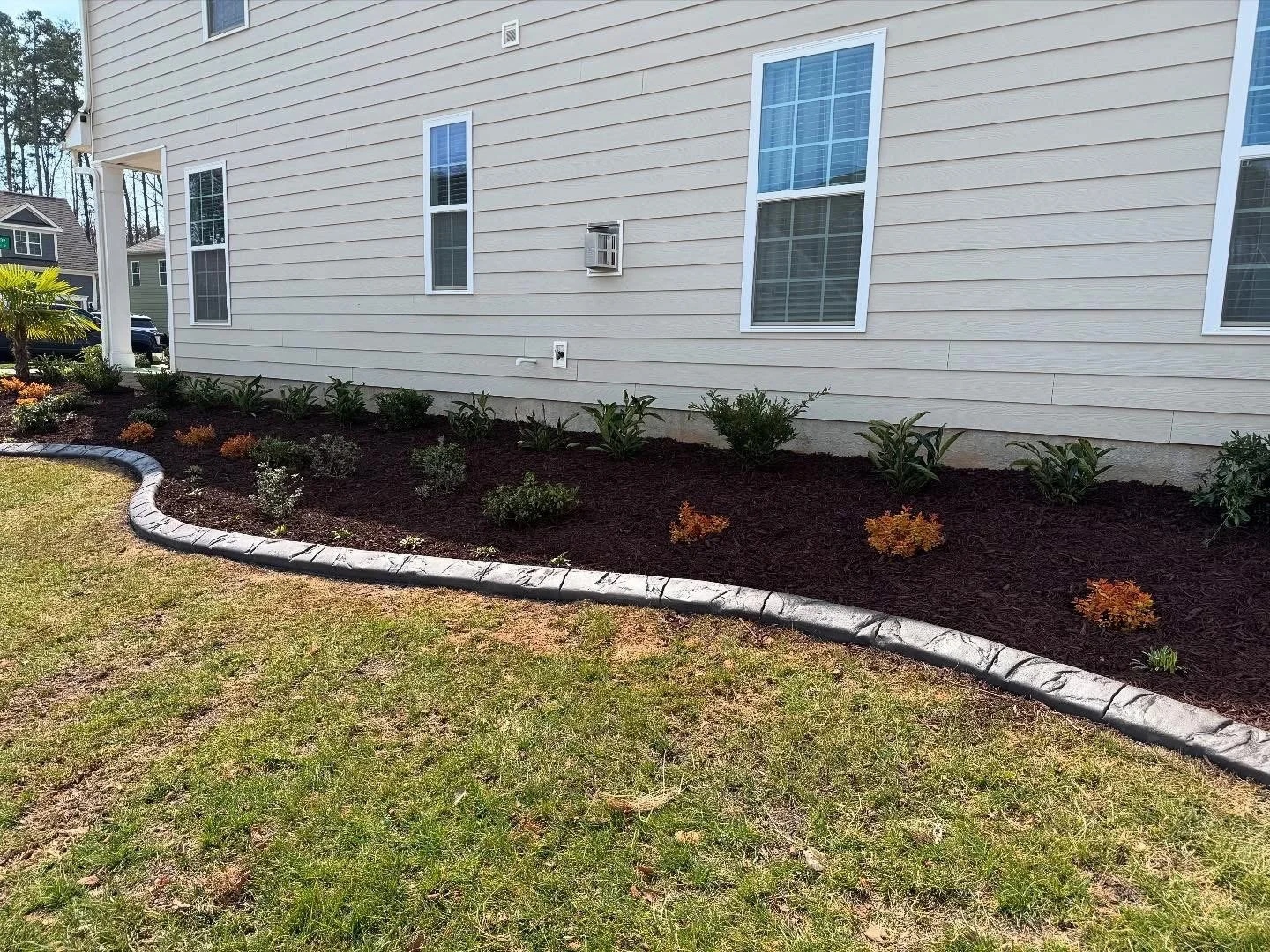 Mulched garden bed with flowers and ornamental plants with stone paver border around the side of a house