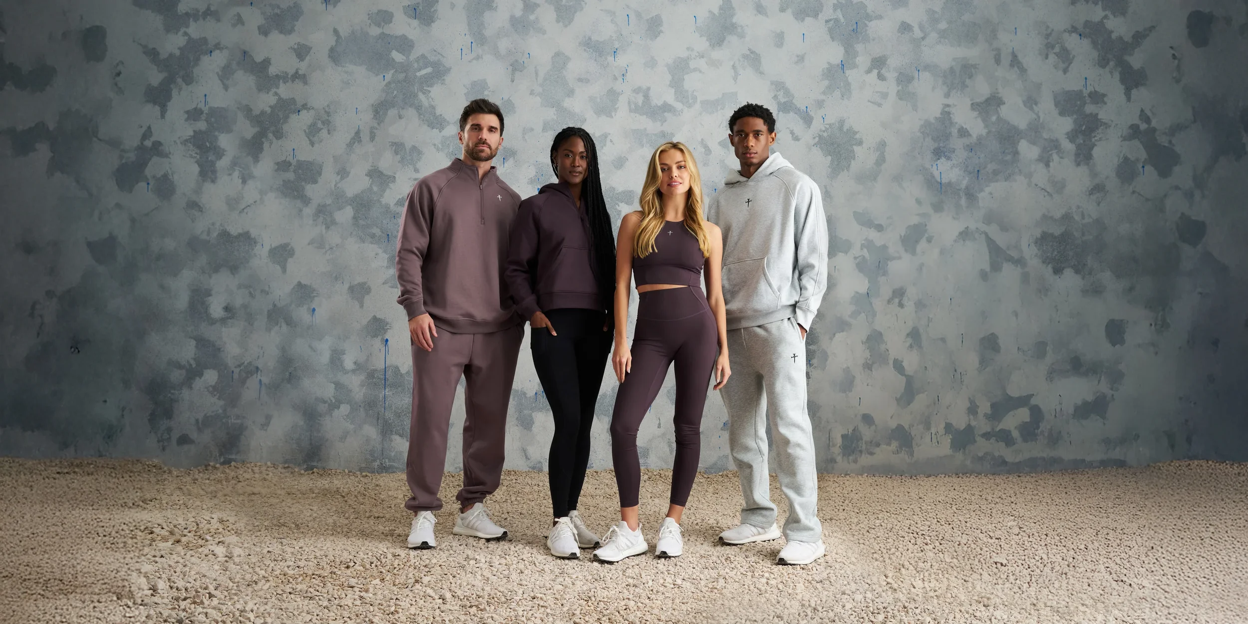 Four diverse people in athletic wear standing in front of a textured gray wall on a gravel floor.