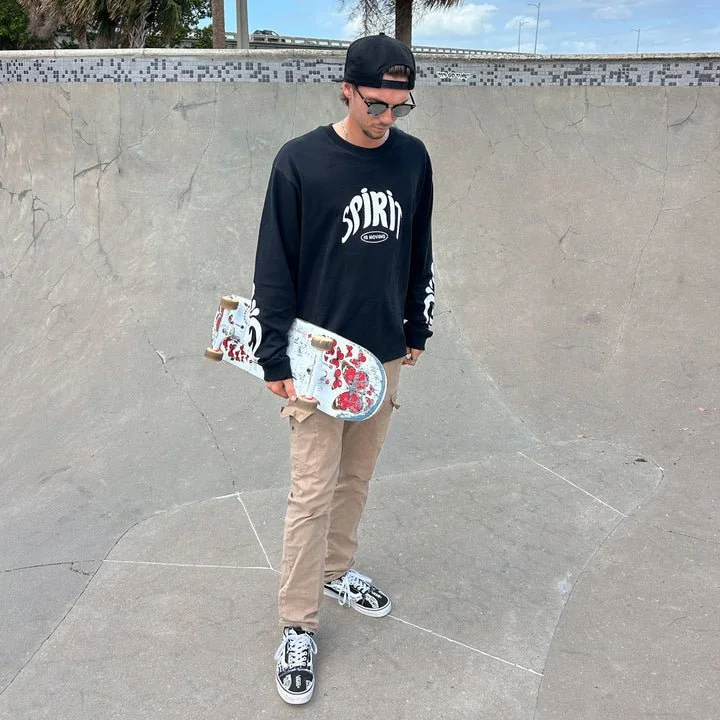 Teenage male skateboarder in black sweatshirt, beige pants, and black and white sneakers at skate park with concrete bowl, holding skateboard with red and white design.