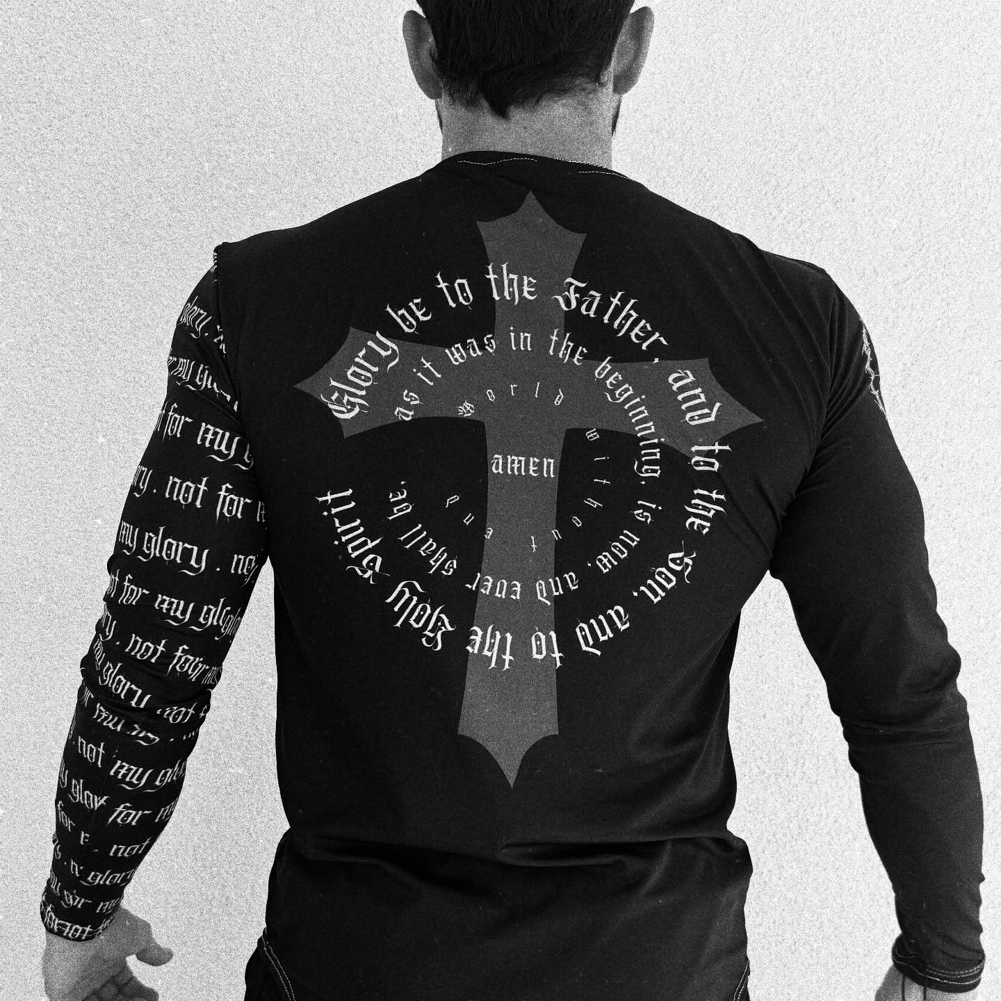 A person with short hair wearing a long-sleeved black shirt with a large cross on the back, surrounded by circular text, and additional text on the sleeves.