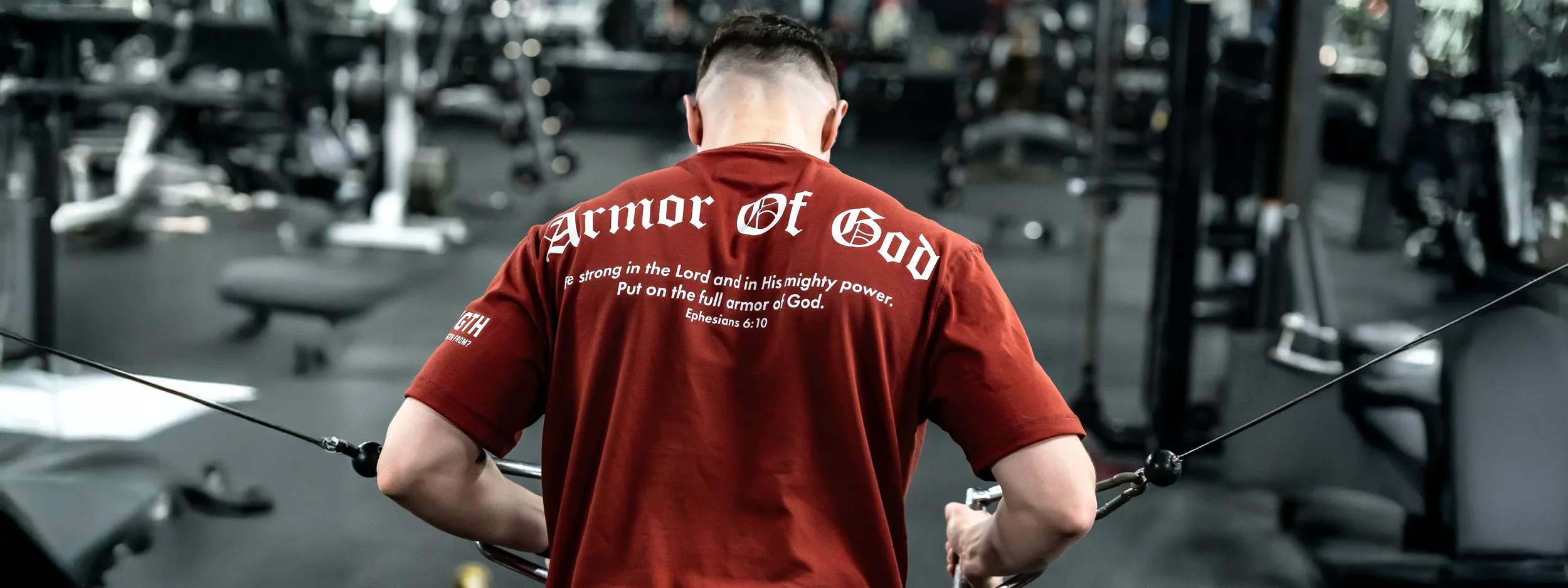 Person in a gym wearing a red T-shirt with a Christian Bible verse on the back, lifting weights or doing pull-down exercises in a fitness center.