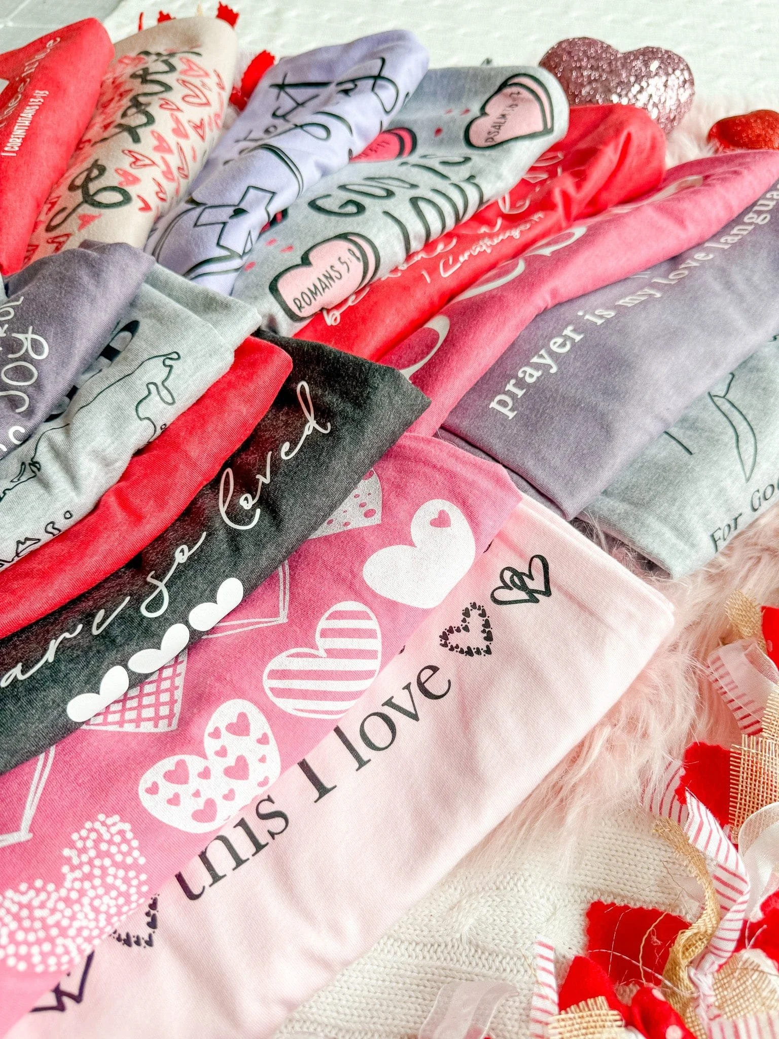 Folded Valentine's Day themed shirts with love and heart designs in pink, red, gray, and black colors, laid on a cream textured fabric with some decorative ribbons and fluffy material at the corner.