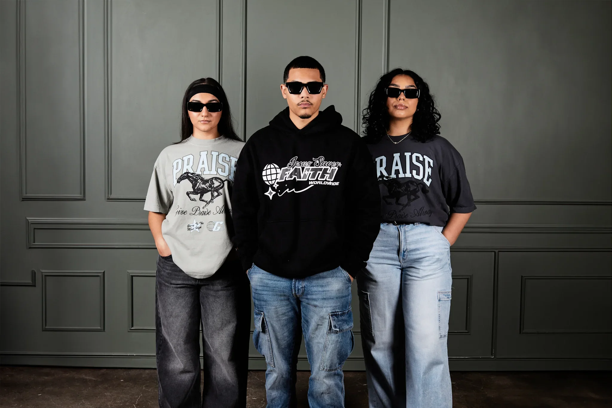 Three young adults standing in front of a gray paneled wall, wearing sunglasses and casual streetwear. The two women are on either side of the man, all with confident expressions.