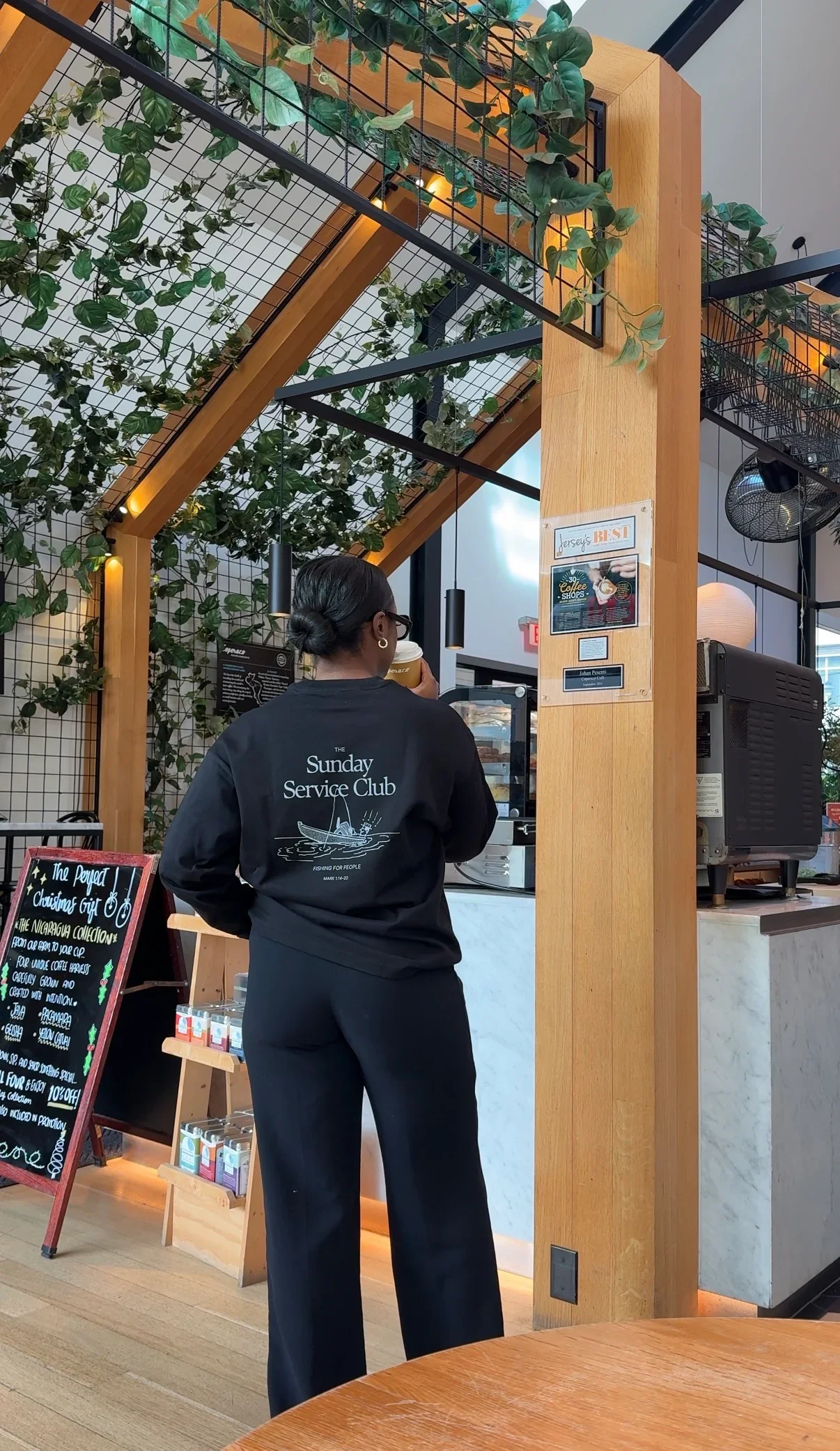 A woman at a coffee shop counter wearing a black sweatshirt with "The Sunday Service Club" logo on the back, holding a disposable coffee cup, with greenery and wooden decor around her.