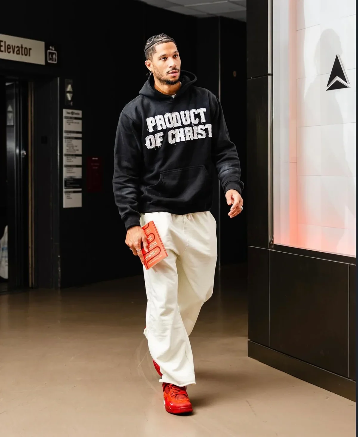 A young man with dark hair and a beard, wearing a black hoodie with the words 'PRODUCT OF CHRIST' on it, white pants, and red sneakers, walking in an indoor setting near an elevator and holding a red book or notebook.