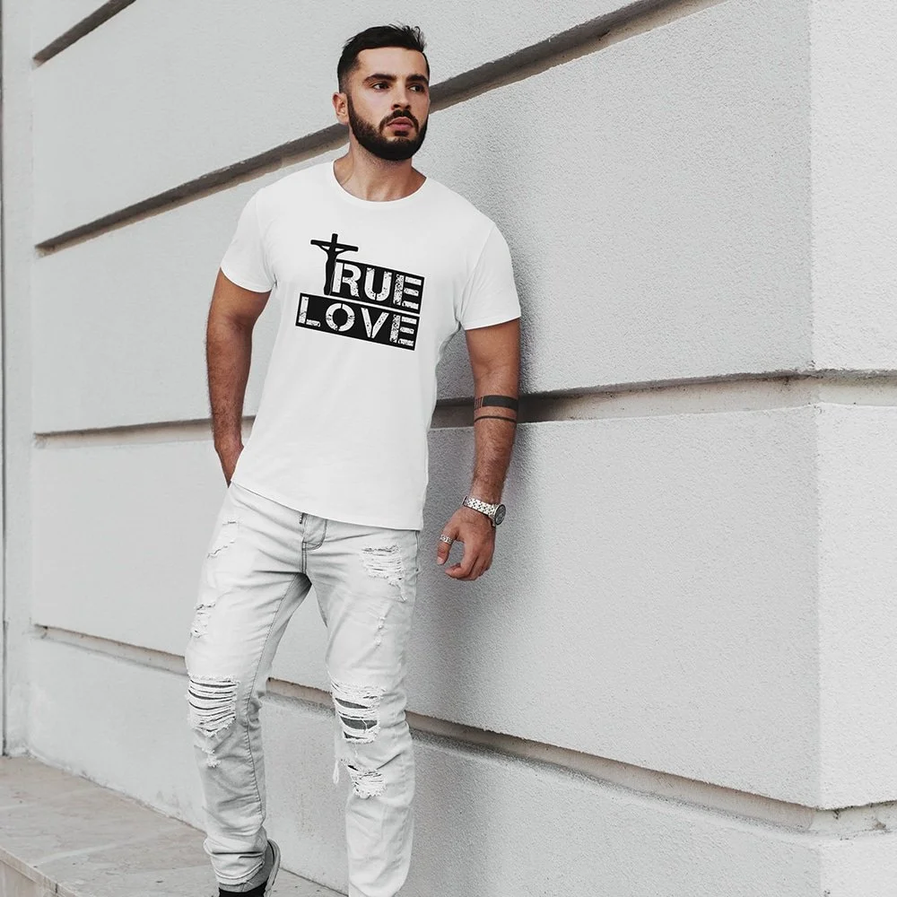 A man with dark hair and a beard standing against a light-colored wall, wearing a white T-shirt with the words "True Love" and a cross on it, distressed white jeans, and a watch.