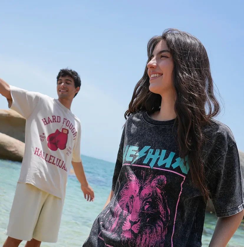 Two young adults smiling and enjoying time at the beach on a sunny day. The woman is in the foreground with long dark hair, wearing a dark graphic t-shirt. The man is in the background with short dark hair, wearing a white t-shirt with boxing gloves graphic and text, near the shoreline with clear blue water.